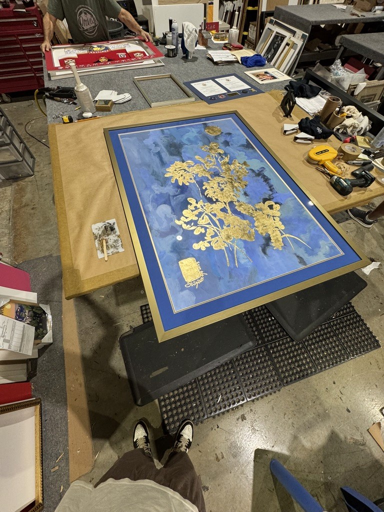 Framing studio table with a gold botanical painting on a blue background being fitted into a frame