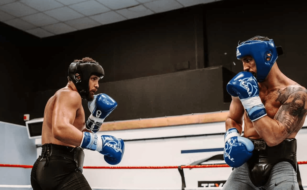 Agron Smakici sparring in headgear practicing defensive footwork and combination punching under coach supervision.