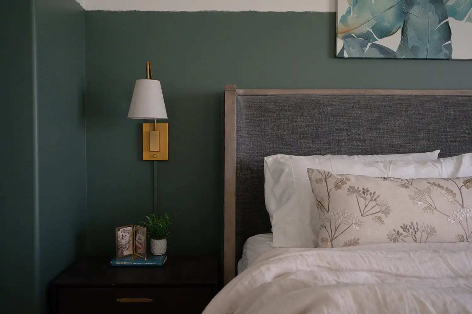 Bedroom interior design featuring a dark forest green two-tone accent wall, linen upholstered headboard, crisp white bedding, floral embroidered accent pillow, brass wall sconce, and botanical watercolour artwork - The Simone Co.