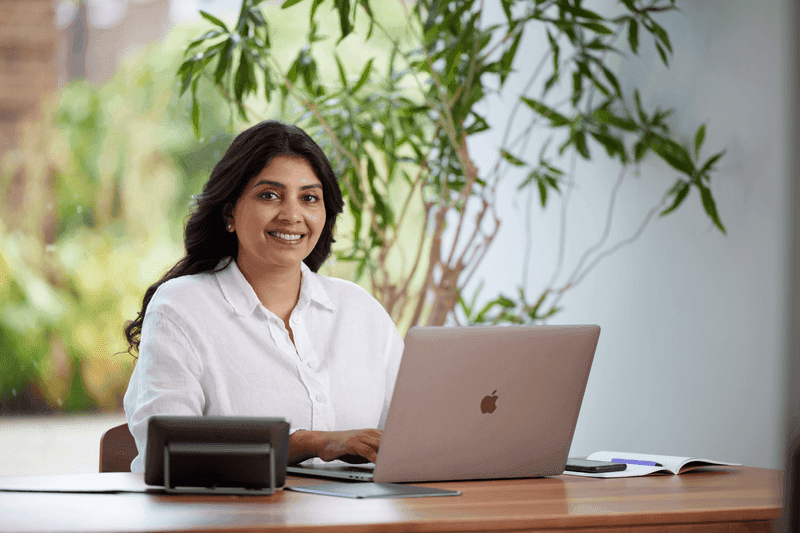 Smiling woman working on a laptop at a wooden desk in a bright office with plants in the background.