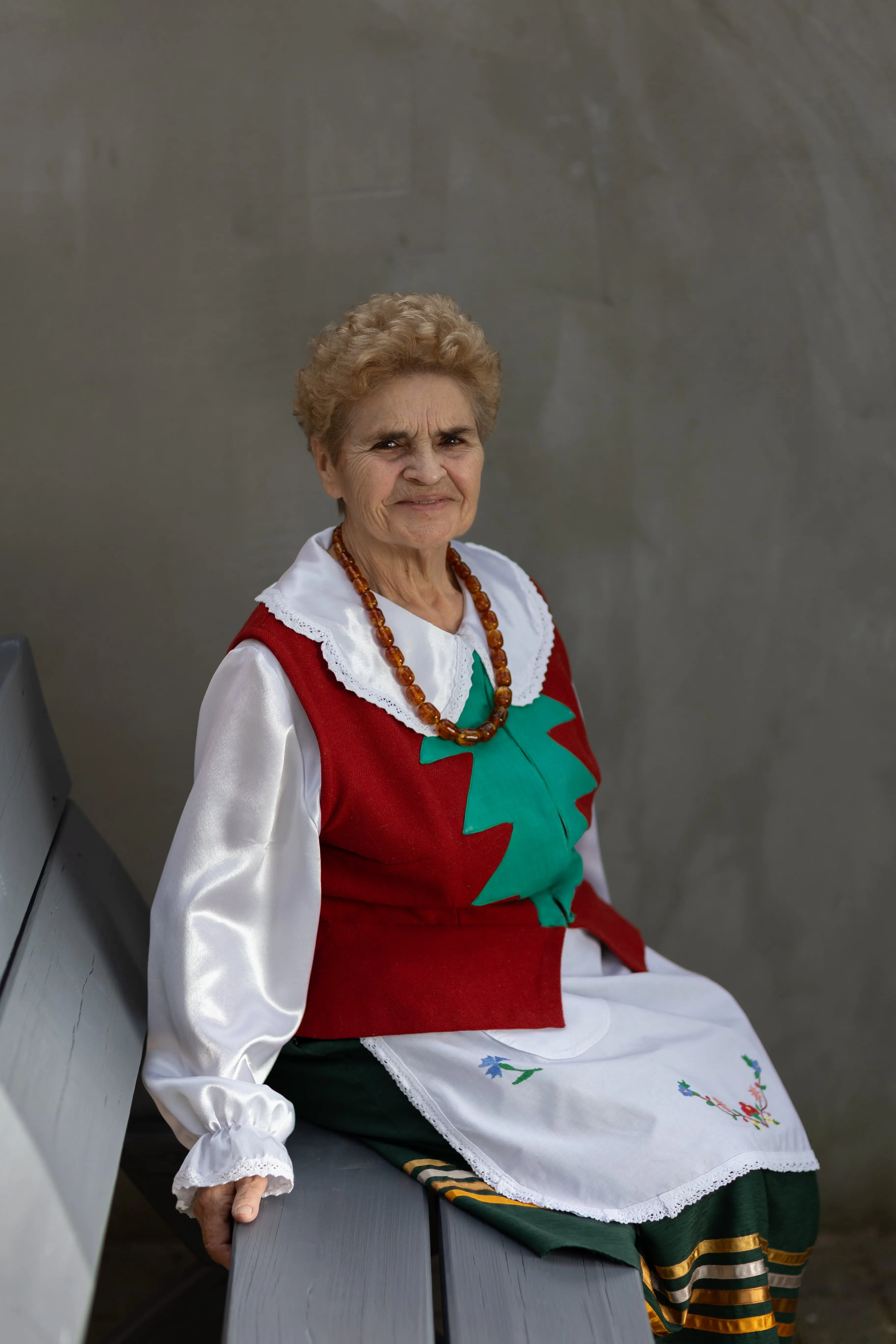 Starsza kobieta w tradycyjnym stroju siedząca na ławce. W tle ściana budynku.