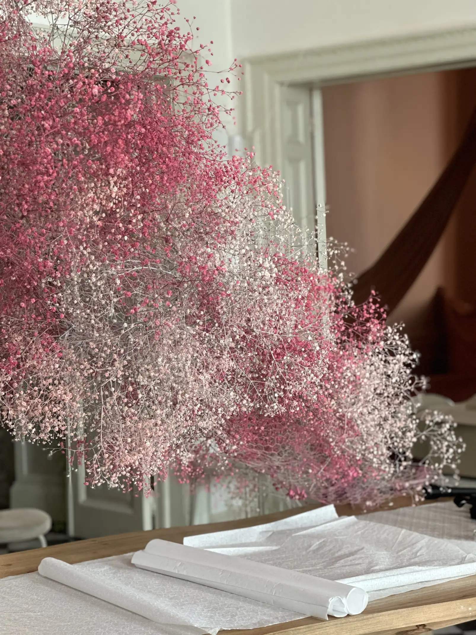 Instalación floral suspendida con ramilletes de flores en tonos rosa y blanco, flotando sobre una mesa de madera cubierta con papel blanco, en una sala luminosa con molduras clásicas y cortinas de fondo.