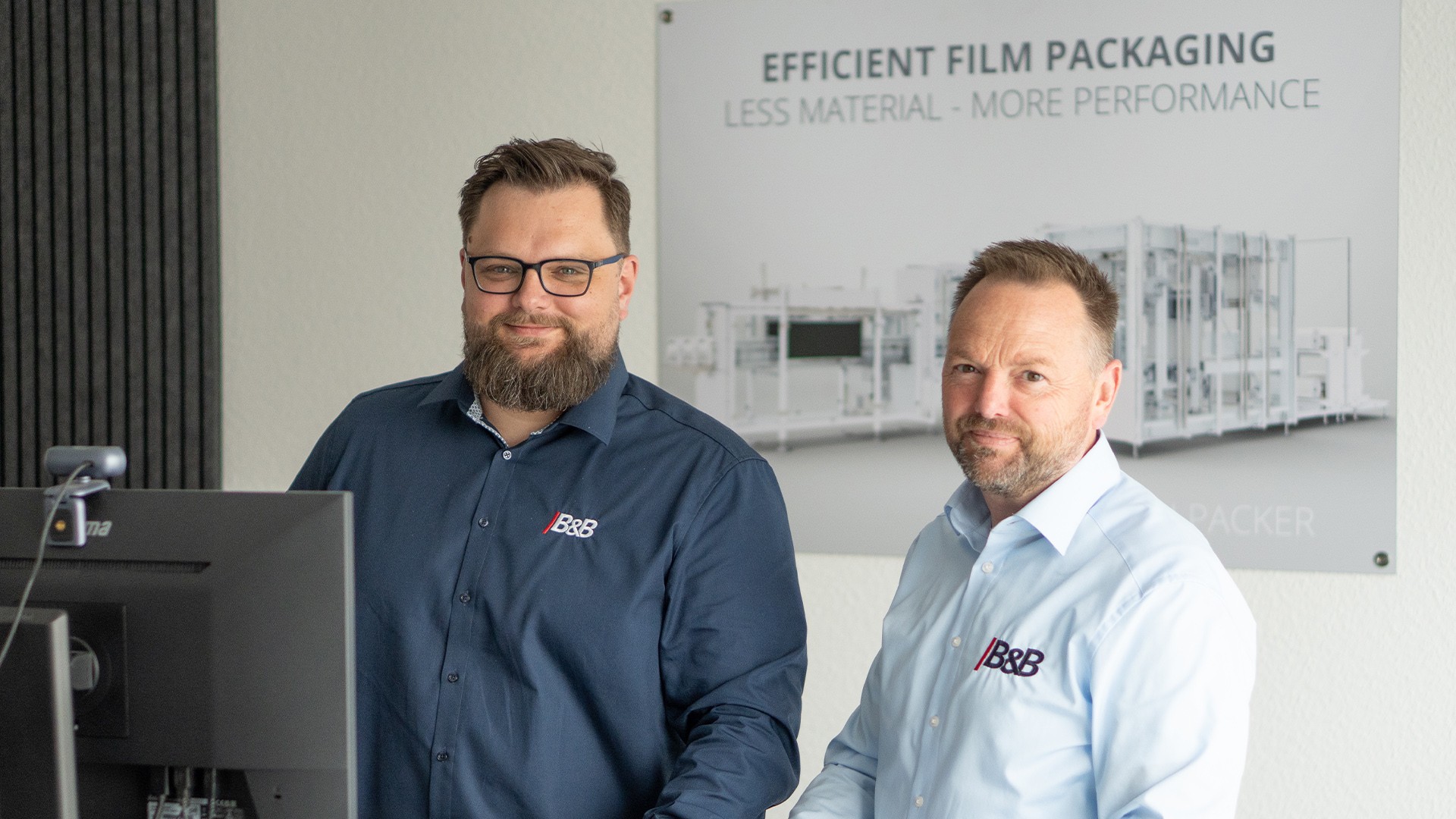 Zwei Männer im Büro, lächelnd in die Kamera. Effizientes Filmverpacken im Hintergrund.