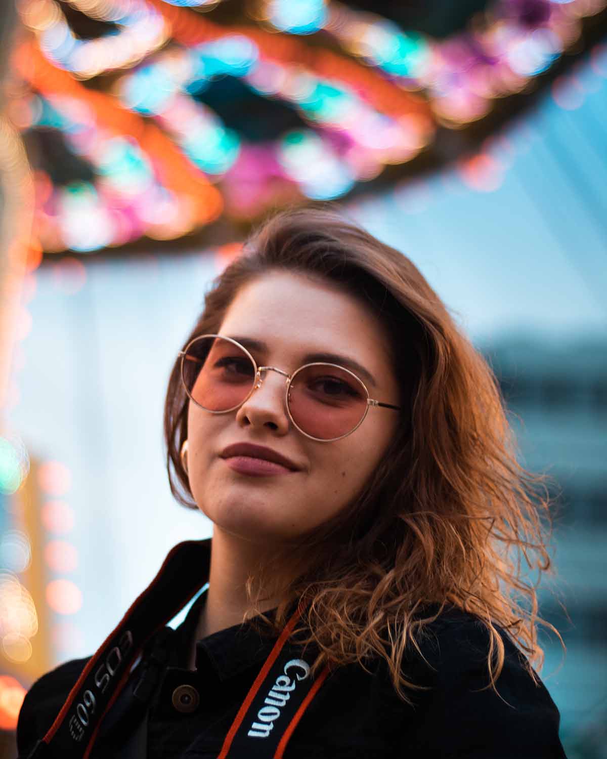 Eine Junge Frau mit getönter Brille schaut in die Kamera sie hat ein Kameraban mit einem Canonlogo um den Hals die der Unschärfe im Hintergrtund sieht man ein Kettencarousell mit Neonlichtern