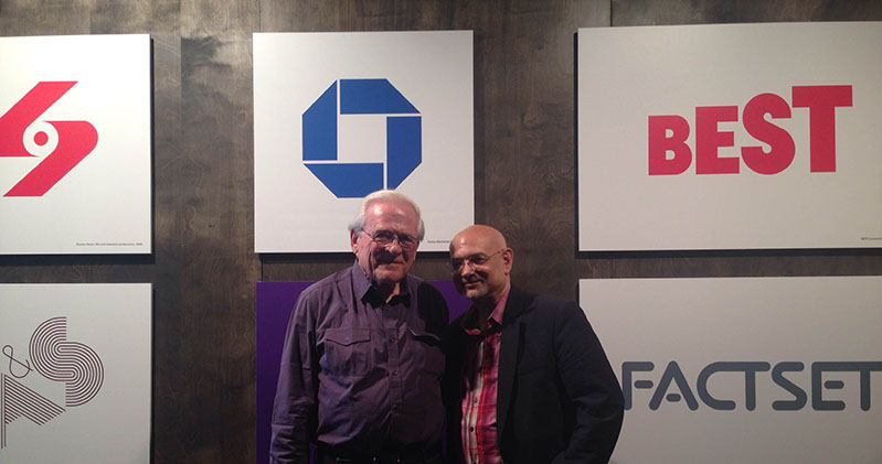 A photo of two men sitting in front of a wall filled with logo placards.