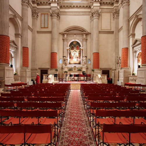 Grand hall with rows of red chairs facing an ornate altar, flanked by tall columns and featuring a decorative red carpet.