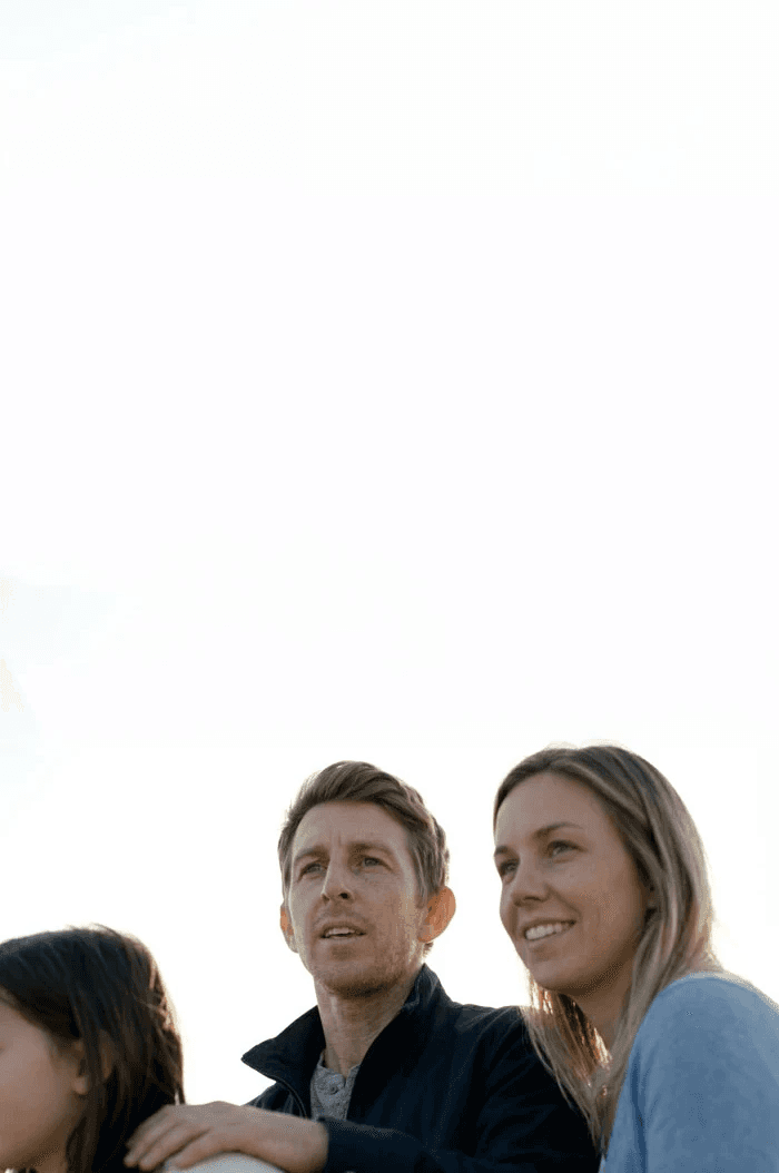 A family stands closely together outdoors under a bright sky, looking content and focused on something in the distance.
