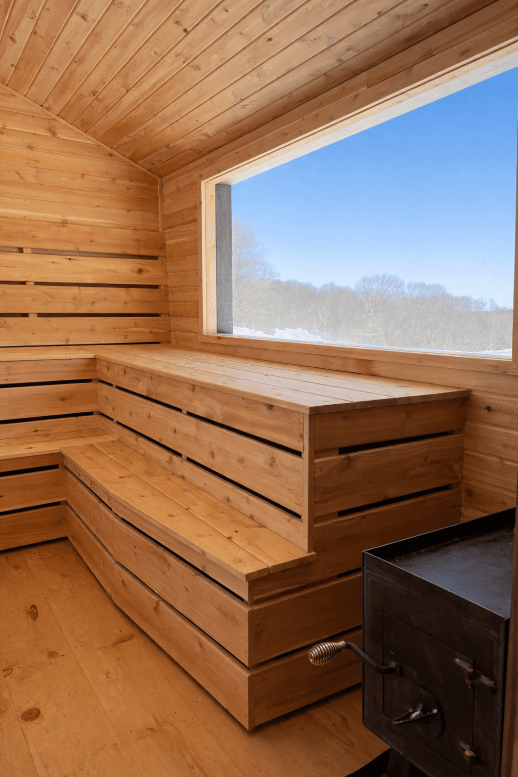 Interior of a cedar wood-fired mobile sauna with tiered benches and panoramic window overlooking a winter landscape.