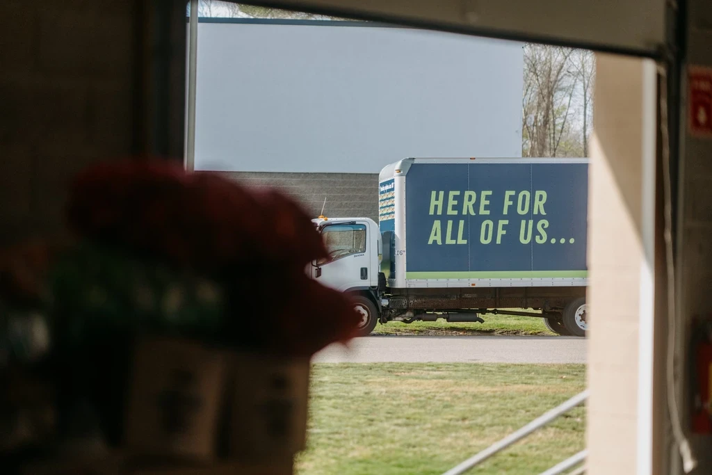 A photograph taken from inside a structure, looking out through an opening or garage door. Outside, a white box truck is parked on a paved area, with a patch of green grass to its left. The side of the truck is dark blue with large, bright green text that reads "HERE FOR ALL OF US...". A large white building is visible in the background. The foreground is blurred, with a red object out of focus on the lower left.