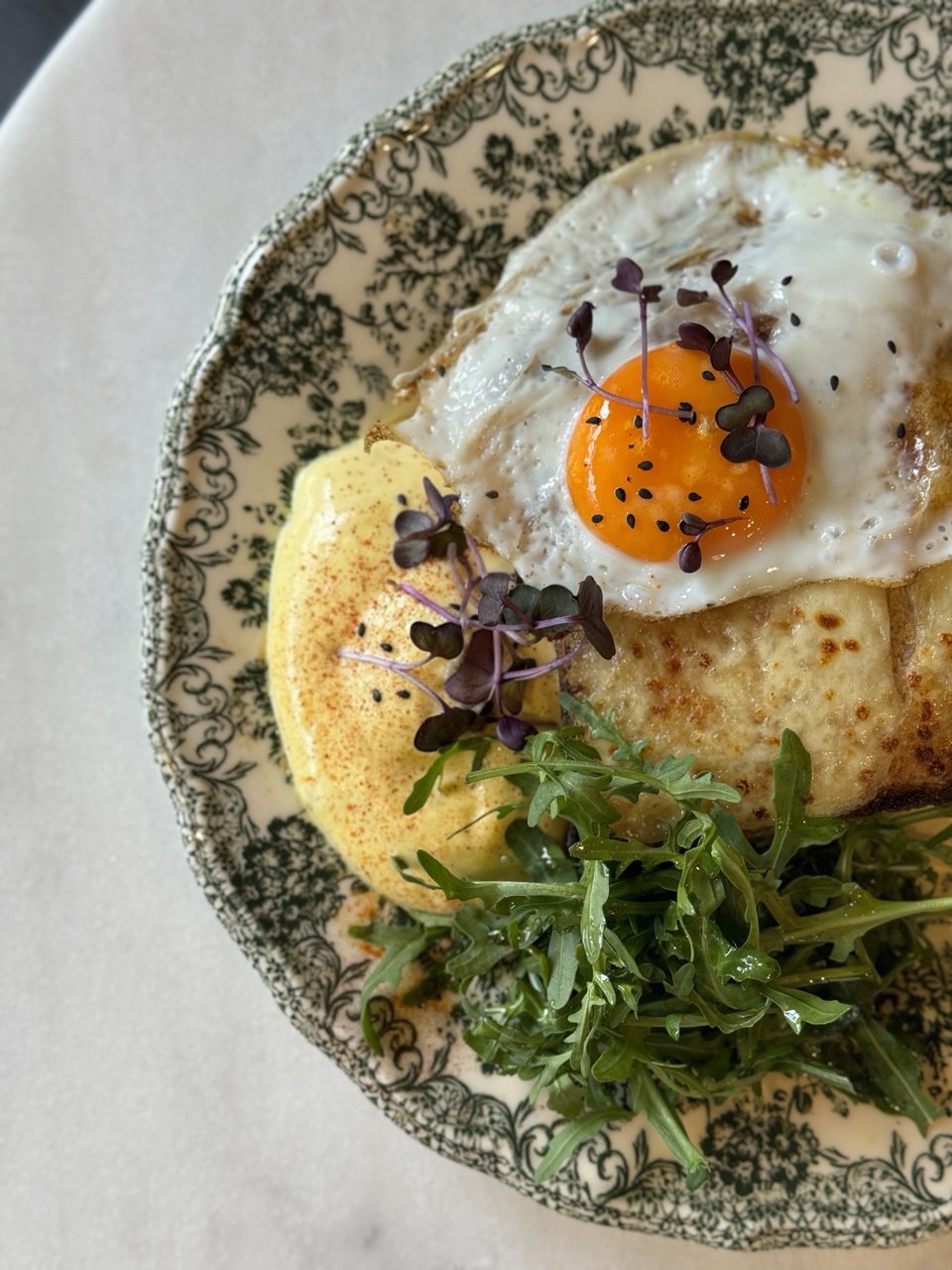 A detailed image shows a savory crepe topped with a sunny-side-up egg garnished with microgreens, accompanied by a side of leafy arugula on a decorative floral-patterned plate, perfect for a gourmet breakfast or brunch setting.
