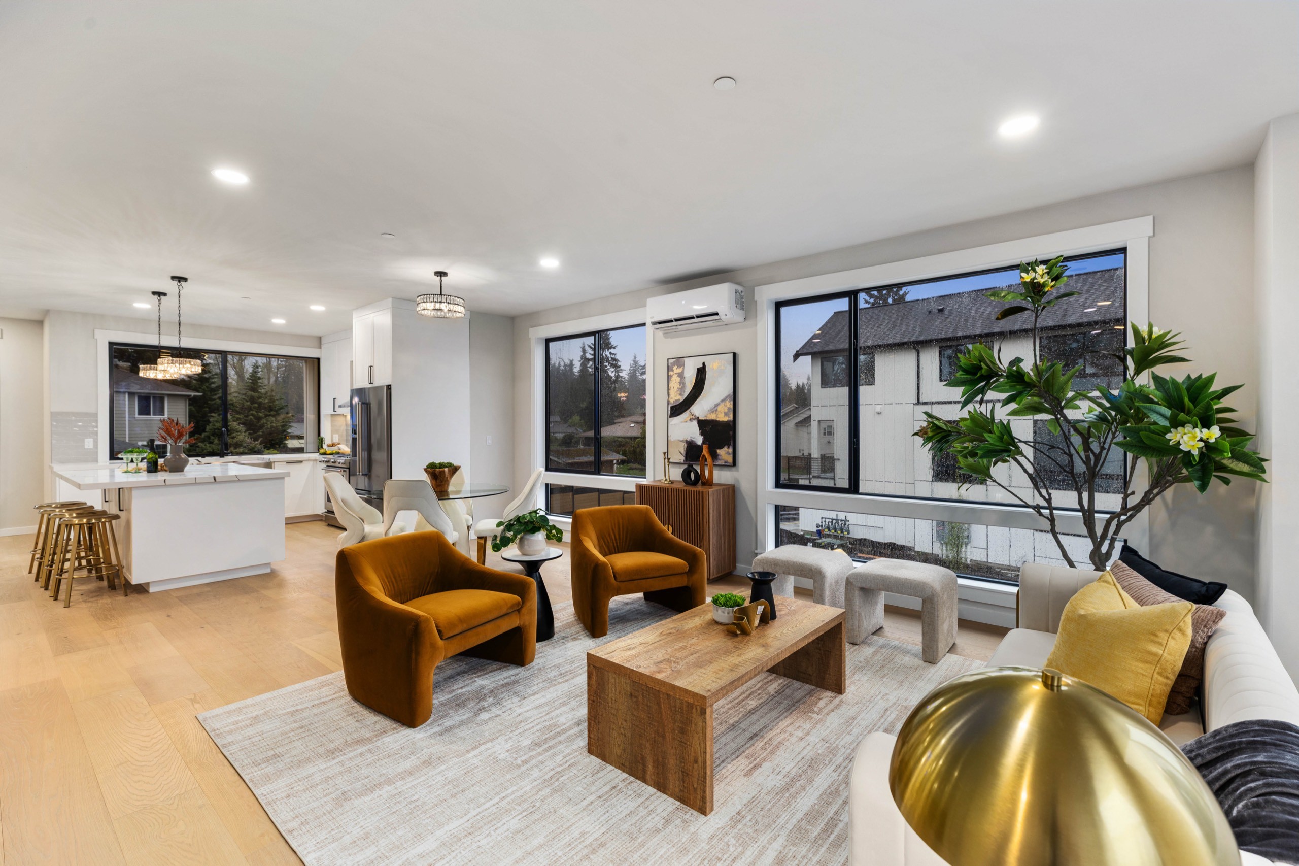 Open-concept luxury living room with kitchen view, amber velvet accent chairs, wood coffee table, oversized windows with garden views, and gold decorative accents