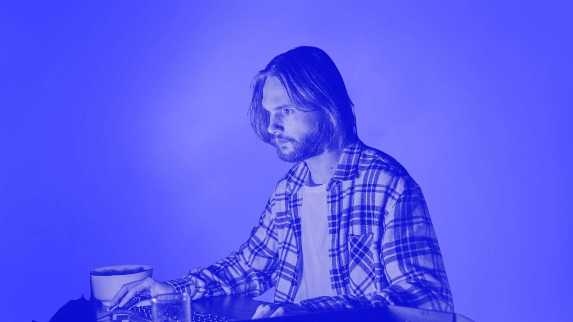 A man with shoulder length hair and a checked shirt sitting at a desk using his computer mouse