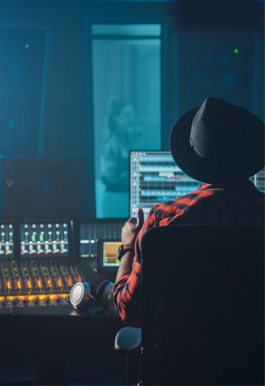 A person in a hat sits in front of a computer in a sound studio, illuminated by blue light in a dark room.