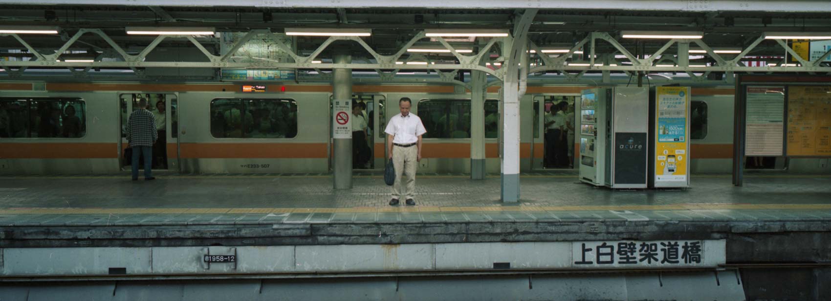 On the platform in the Japanese metro a man is waiting for a train looking down at the ground, horizontal art captured on Hasselblad XPan, alone by himself on the platform in Tokyo Japan