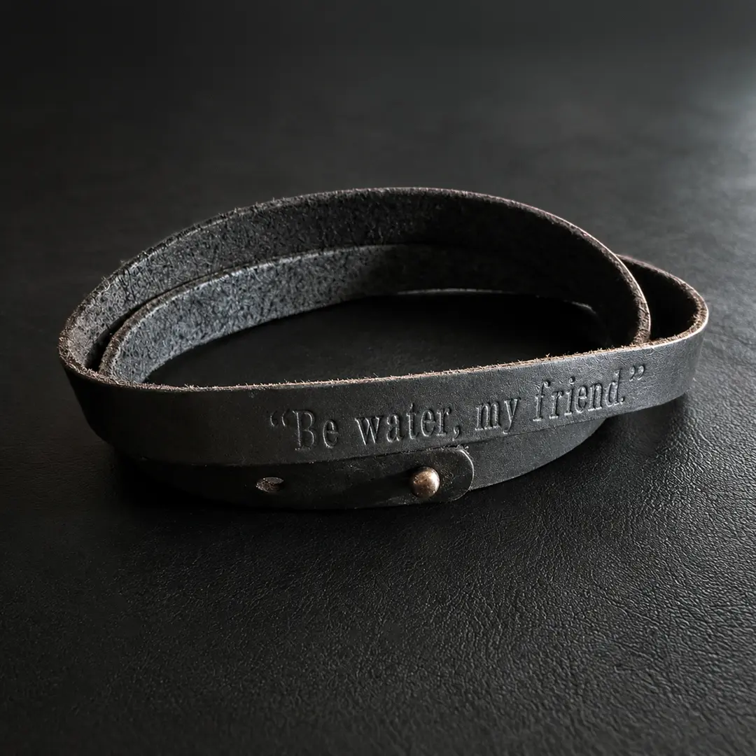 Black leather wrist wrap engraved with “Be water, my friend.” resting on a dark desk, with soft side lighting highlighting the lettering and worn texture.