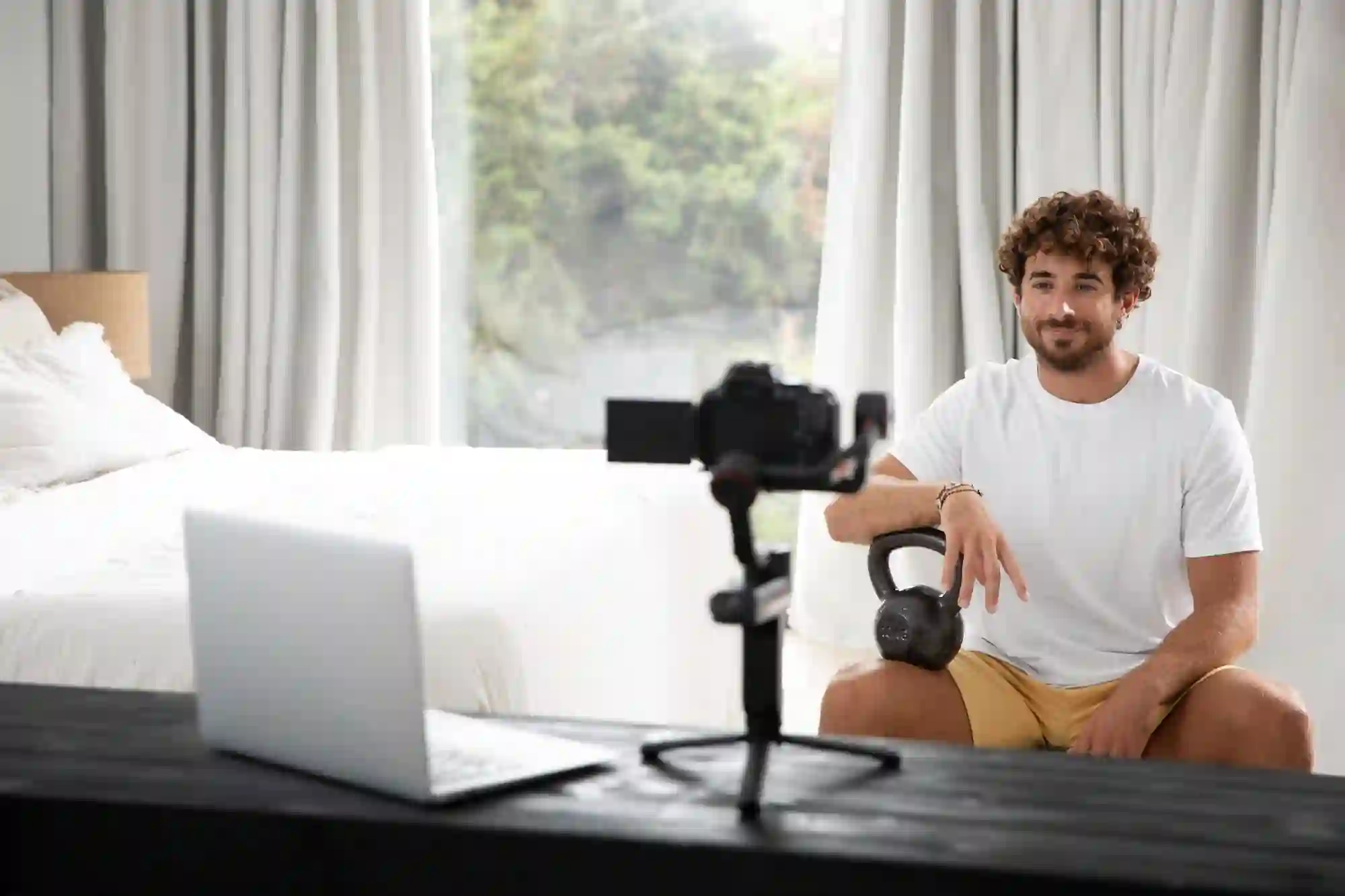 A smiling man filming a fitness video at home with a kettlebell, camera, and laptop.