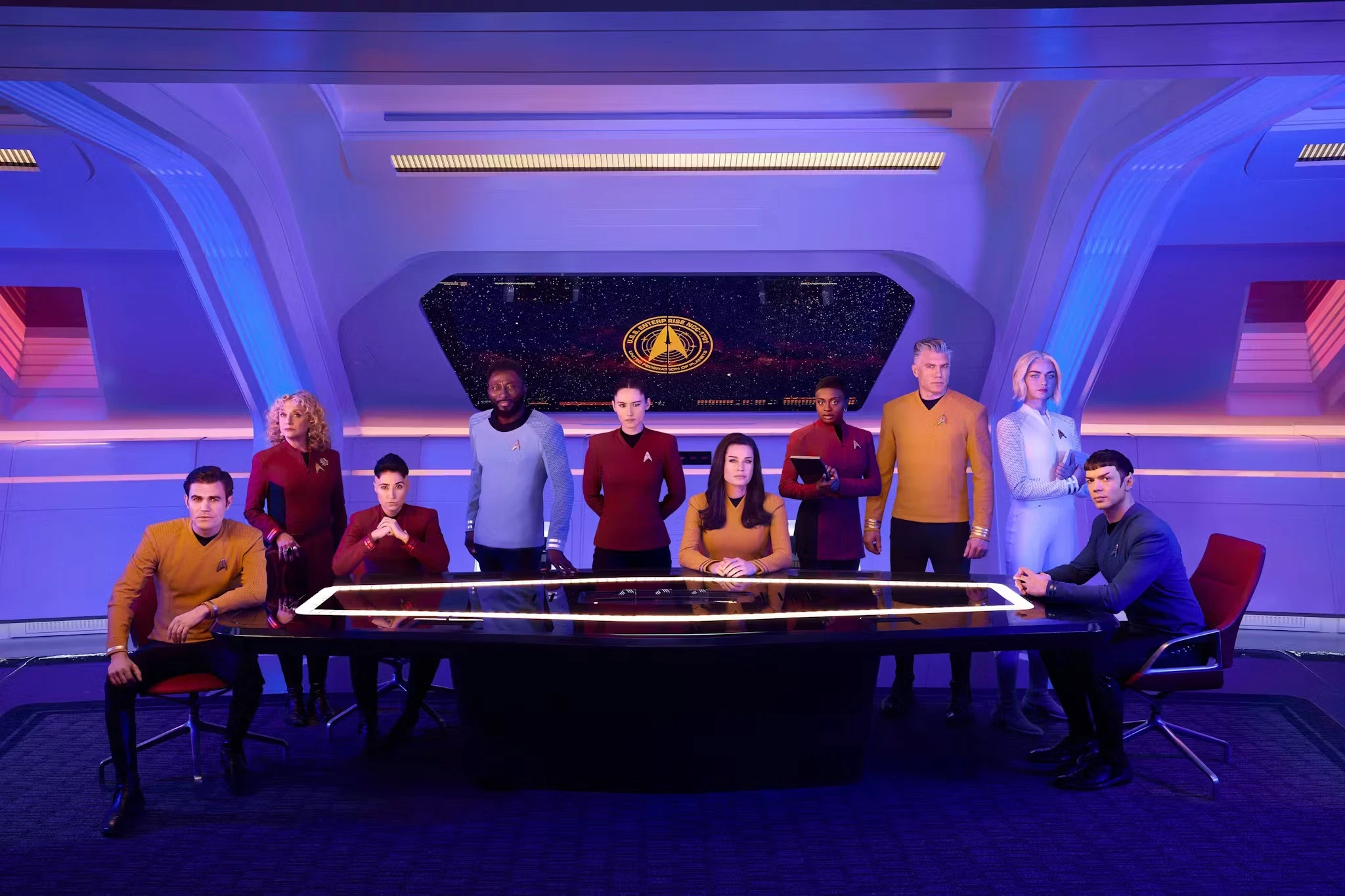 A group of uniformed people are gathered in a futuristic room, sitting and standing around a glowing table. A large window displays a starry view with a circular emblem. The room is bathed in blue and red lighting.