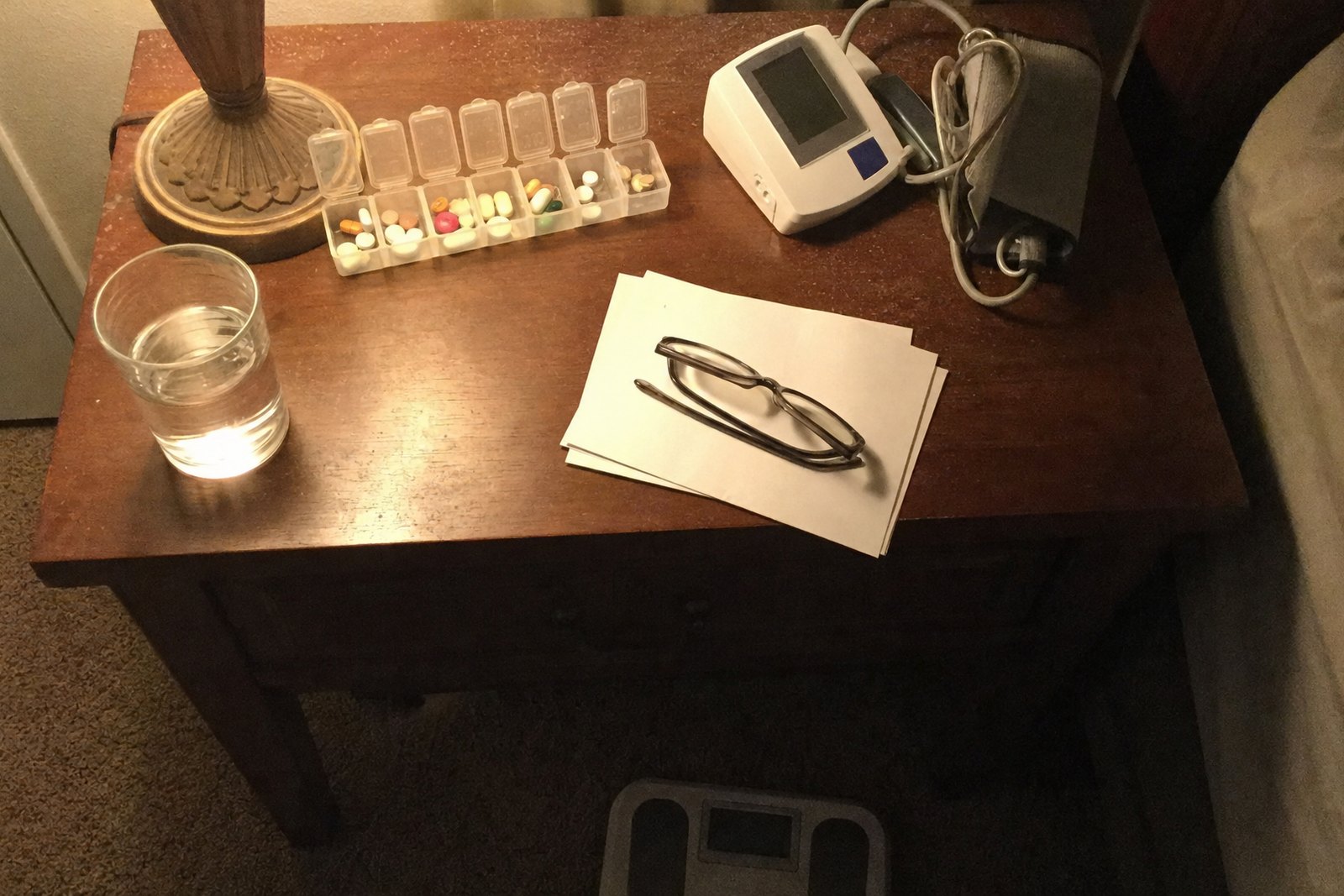 A weekly pill organizer with its lids open beside a home blood pressure cuff, reading glasses on a stack of blank papers, and a glass of water on a bedside table at night.