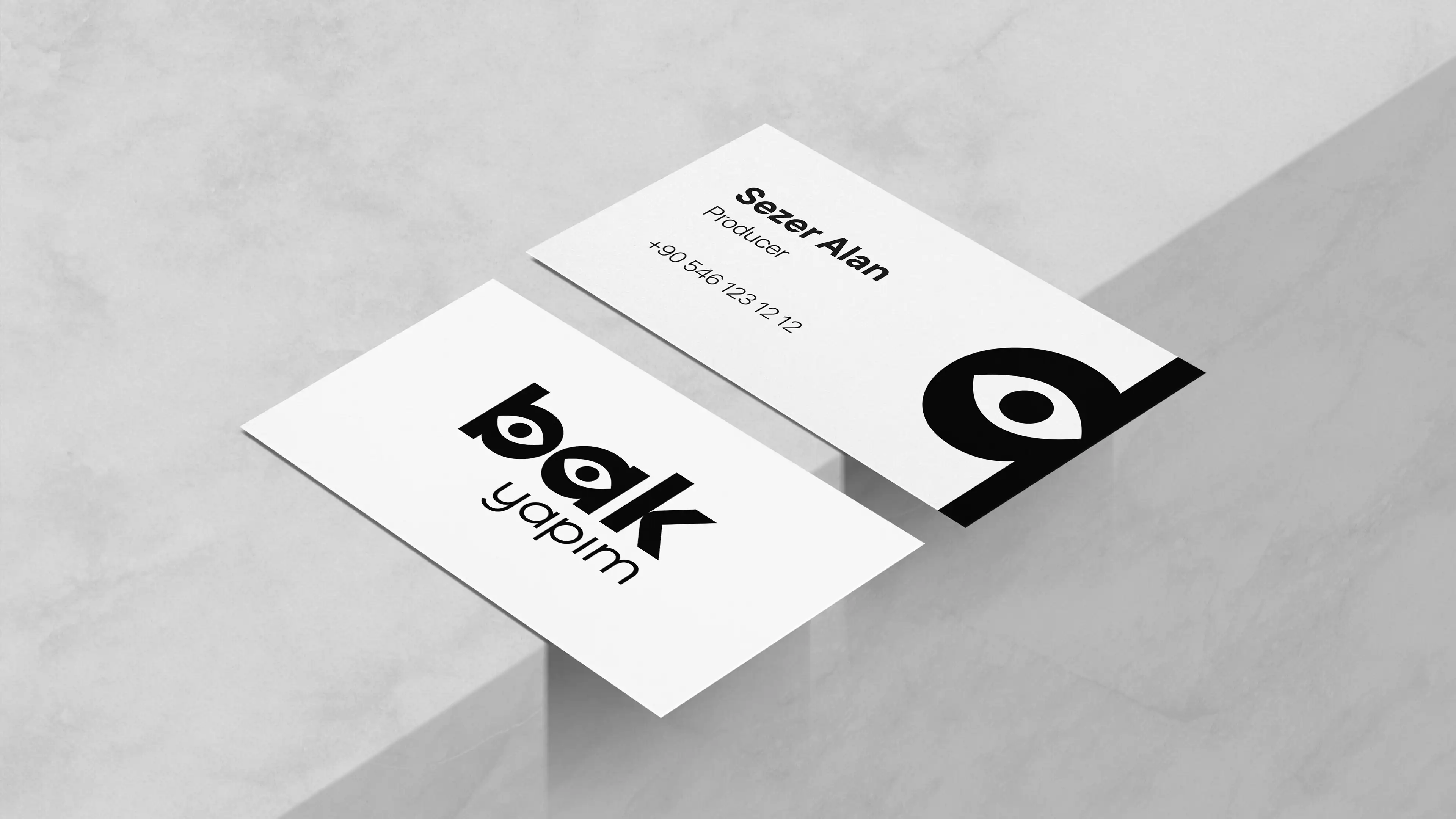 Professional white business cards for producer Sezer Alan from Bak Yapım resting on a minimalist grey concrete architectural surface.
