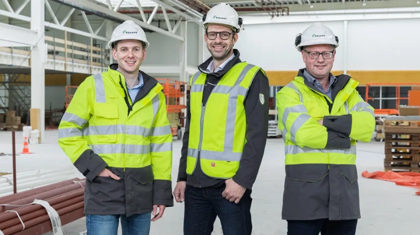 Three people wearing safety helmets and high-visibility jackets stand confidently inside a large, industrial warehouse under construction, surrounded by building materials and equipment.