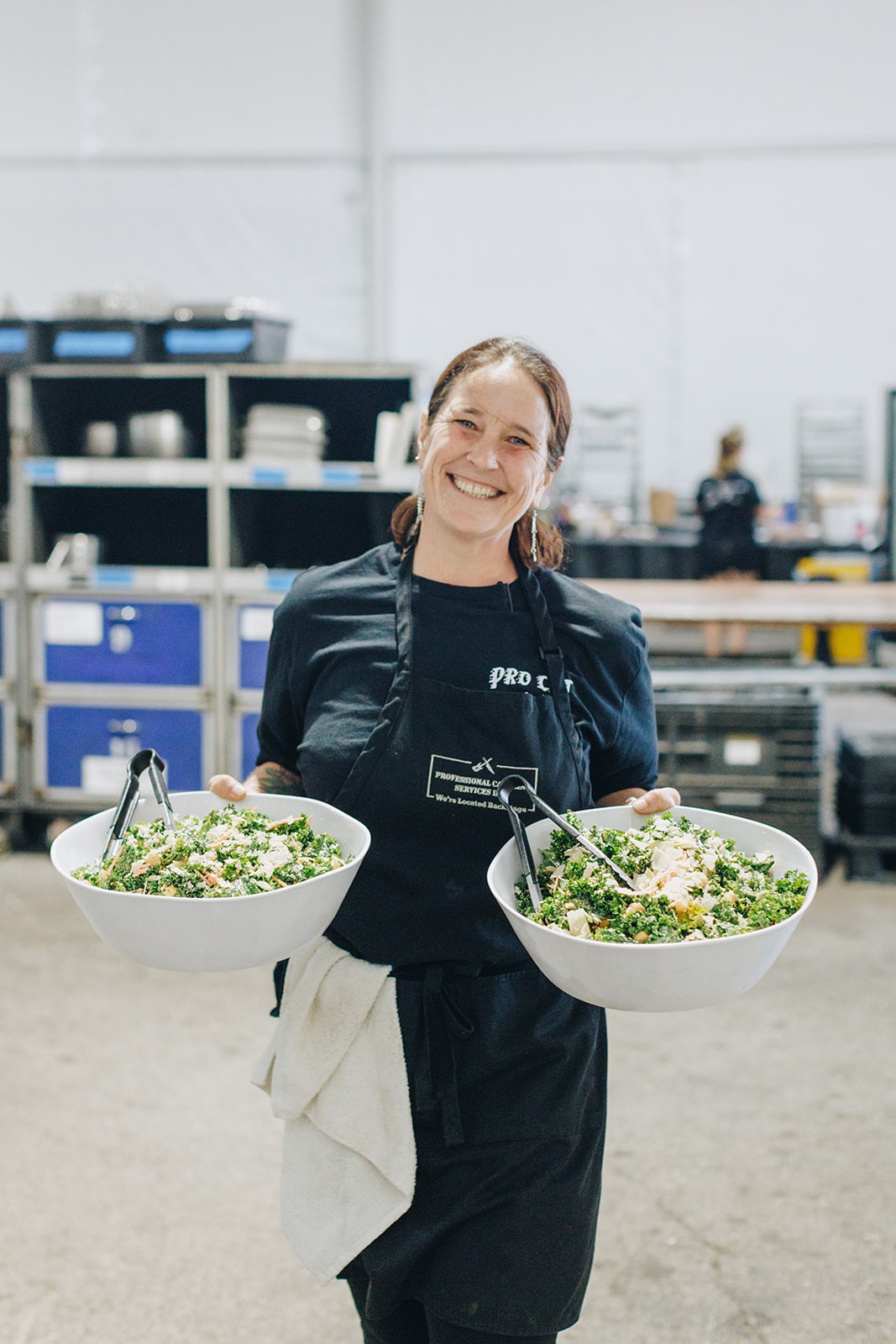A person wearing a black jacket holds two plates of food, smiling in a kitchen setting.