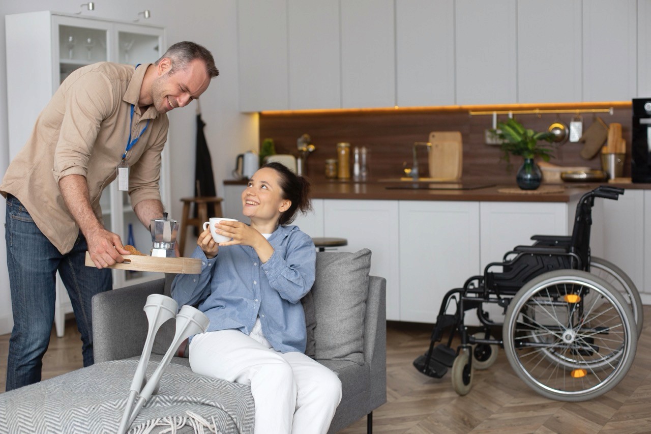 A smiling man serves a drink on a tray to a woman sitting on a couch with crutches beside her. A wheelchair is visible in the background in a modern kitchen and living area.