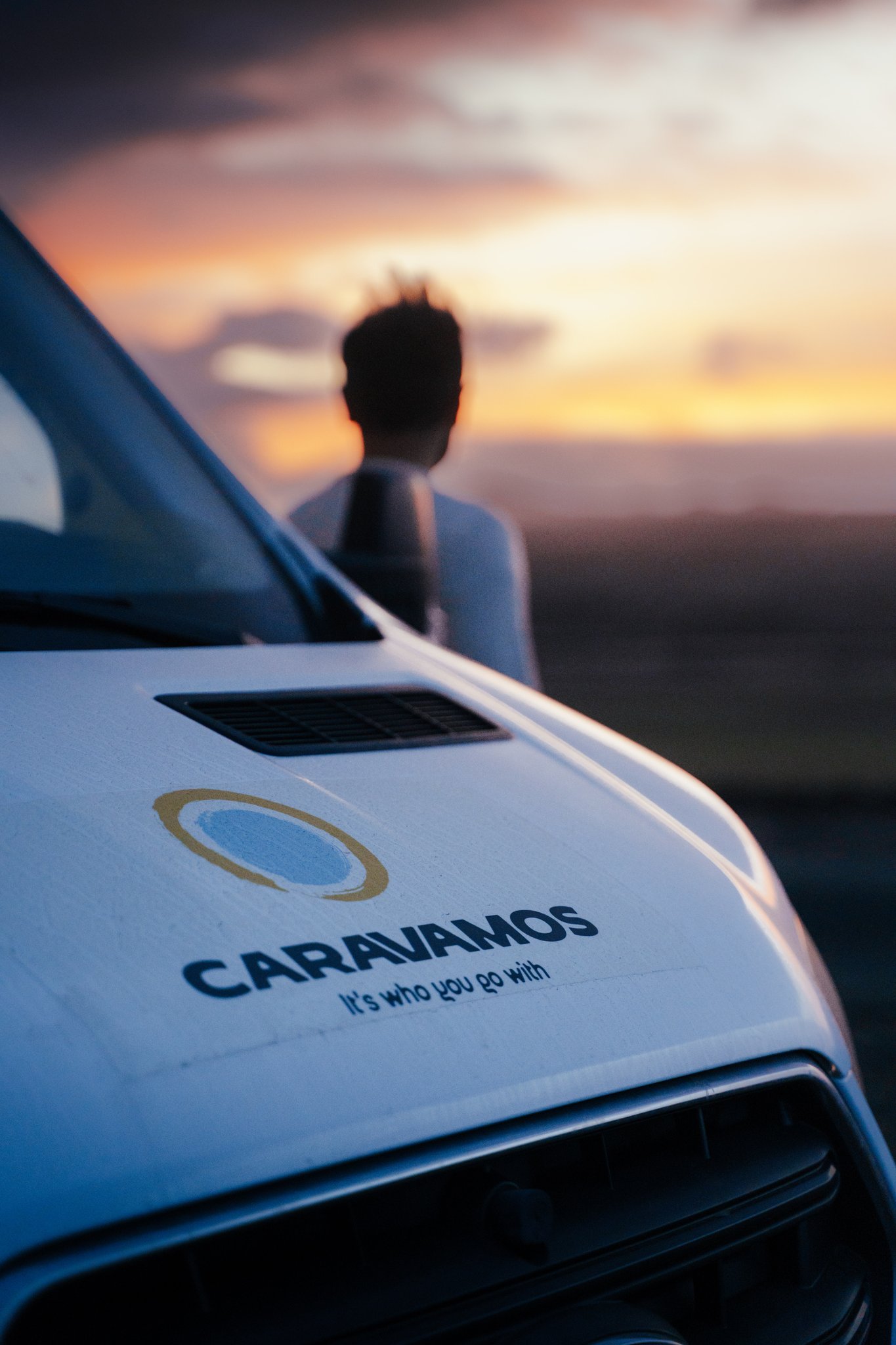A person standing in the background beside a vehicle, with a sunset sky and the vehicle's logo in focus.