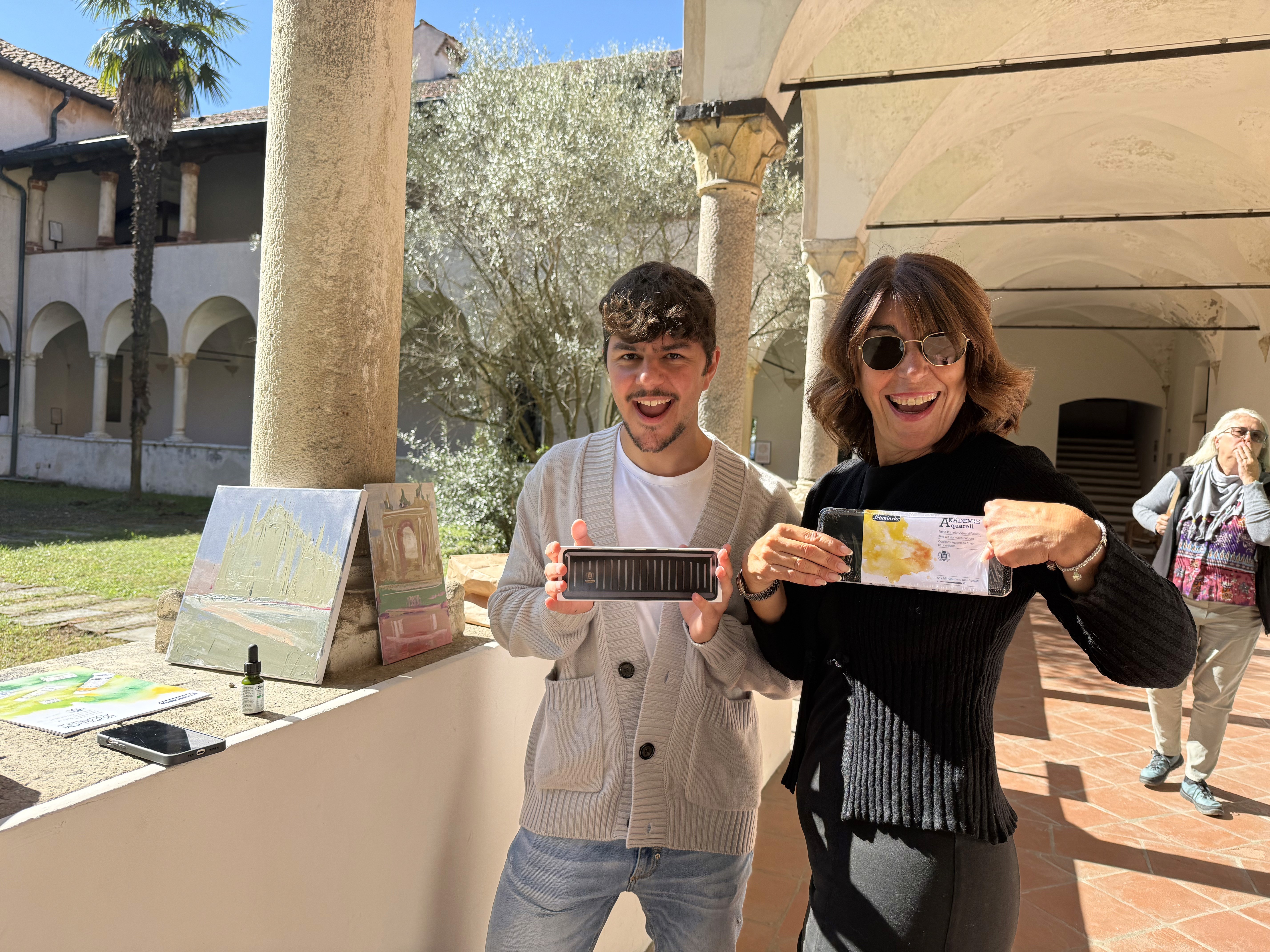 Two people smile and hold up a rectangular object and a card in a sunlit veranda with columns and artwork in the background, inside a historic building.