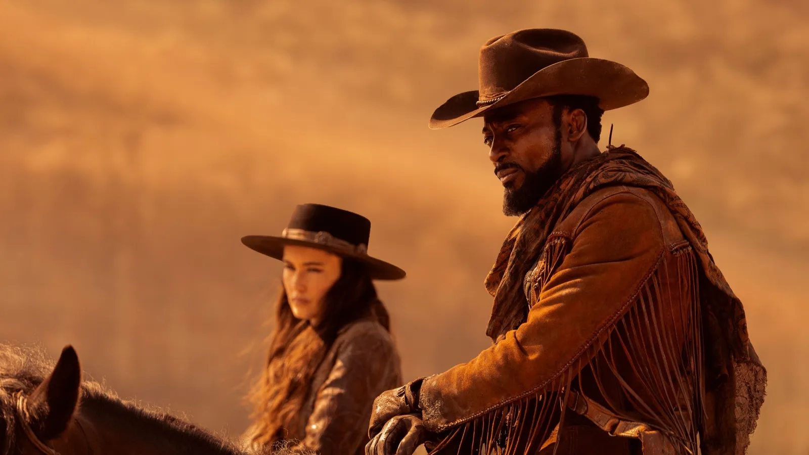 A man and woman in traditional Western cowboy attire, featuring hats and fringe jackets, ride horses against a backdrop of a sunlit, dusty desert landscape, embodying classic Wild West imagery.