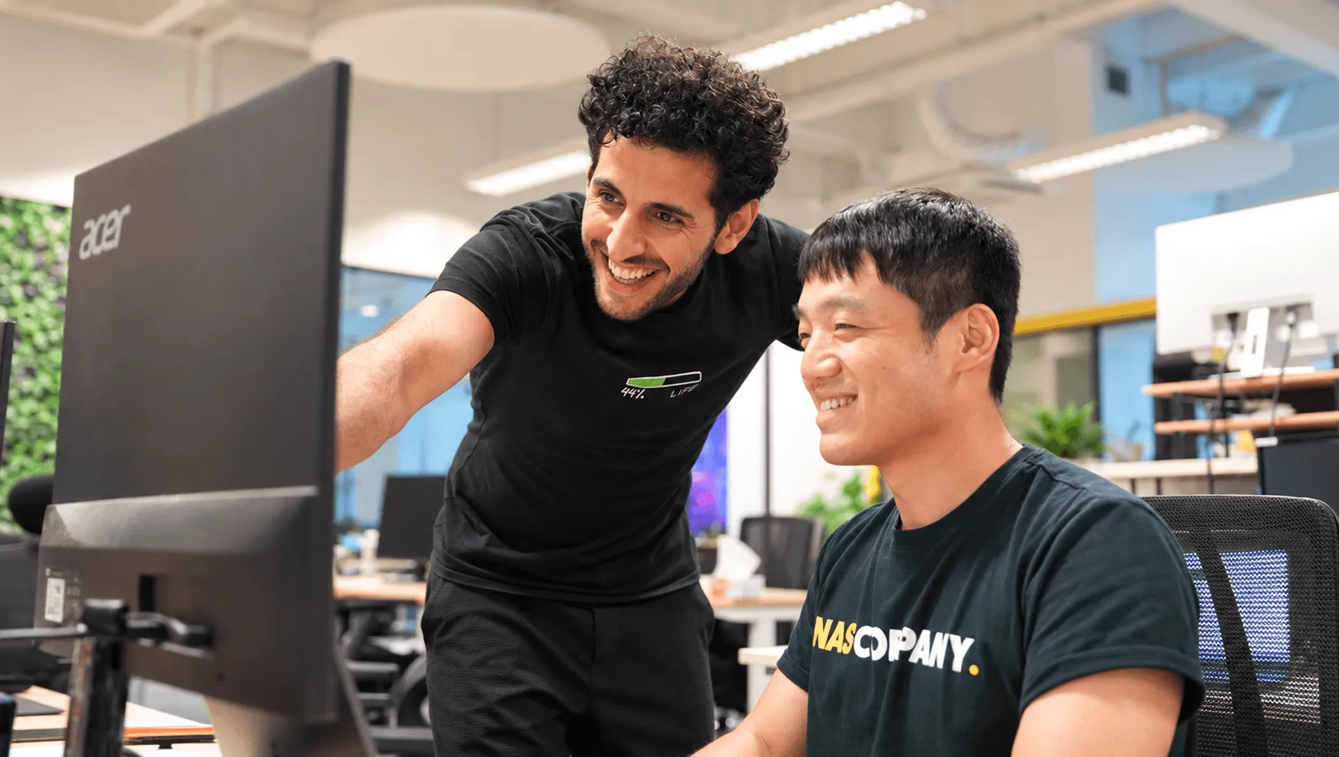 Two Nas.com team members smiling while reviewing work on a monitor