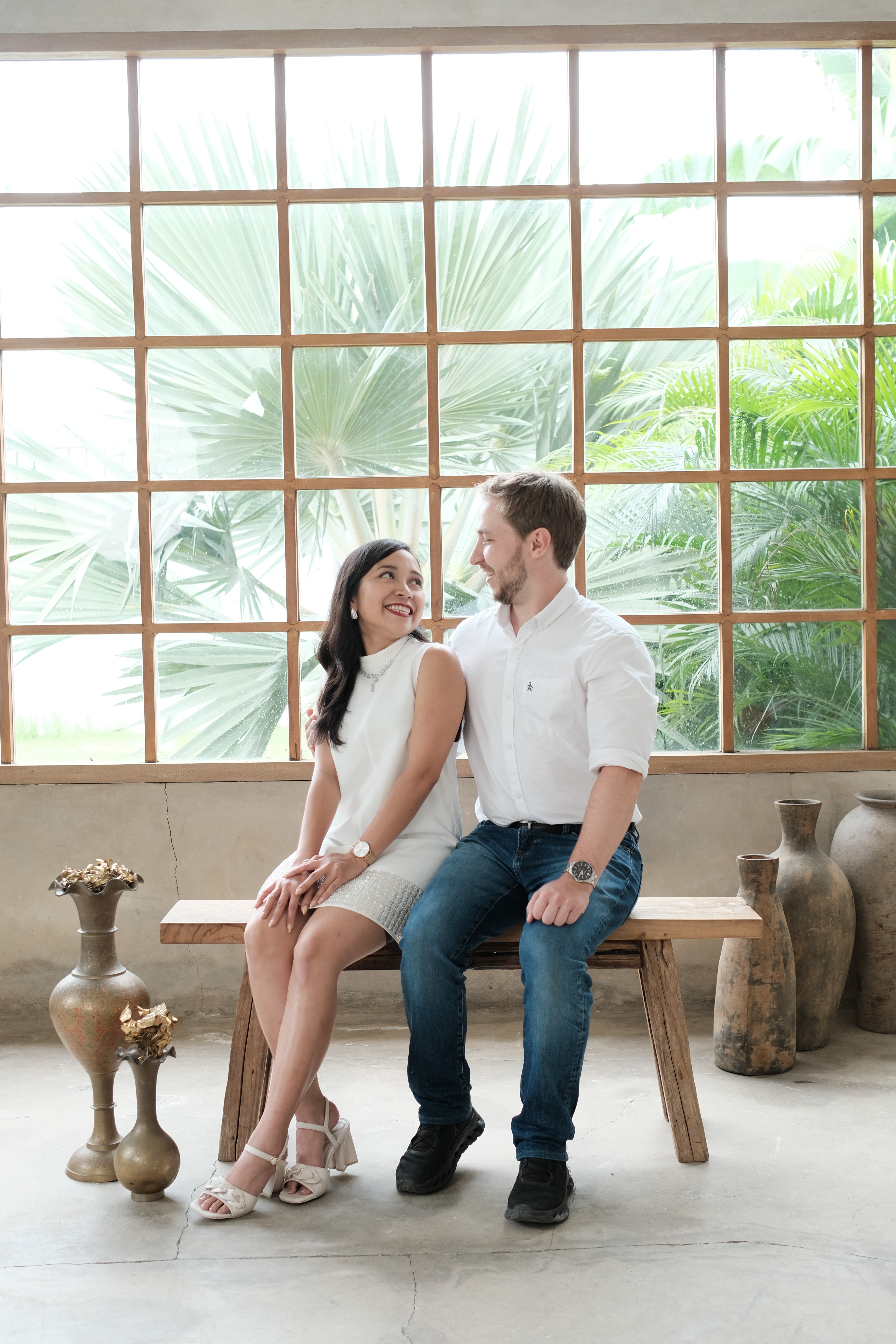 A bride and groom hold hands tenderly, the bride's bouquet of dried flowers hanging low. They stand in a softly lit, elegant interior, conveying romance.