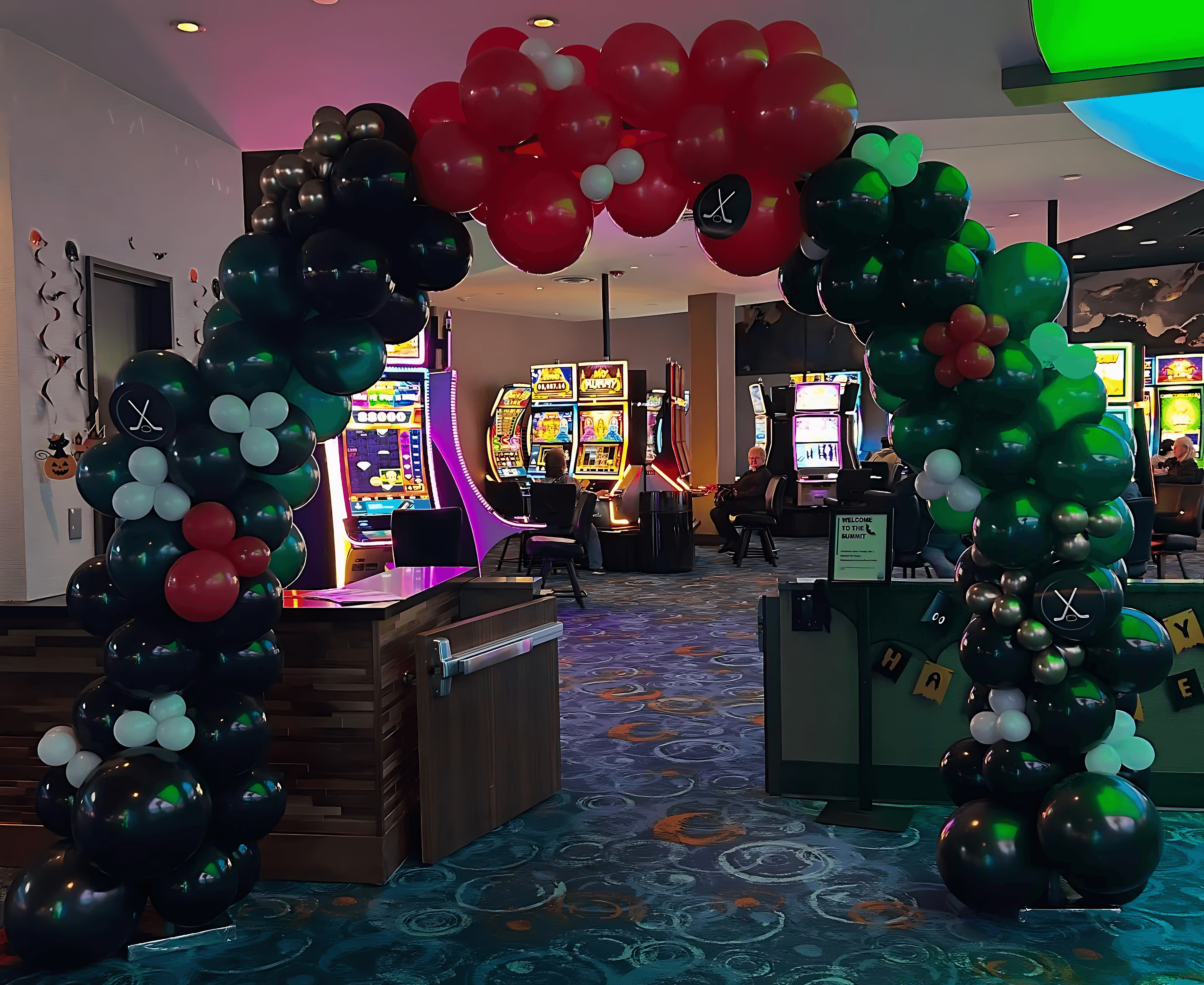 A festive balloon arch in red, green, and black frames the entrance of a lively casino, with slot machines and vibrant decor enhancing the colourful atmosphere.