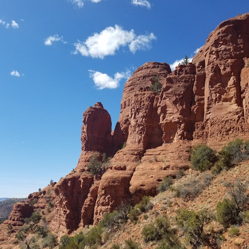 Червени скални образувания с оскъдна растителност под ясно, синьо небе с няколко бели облака.