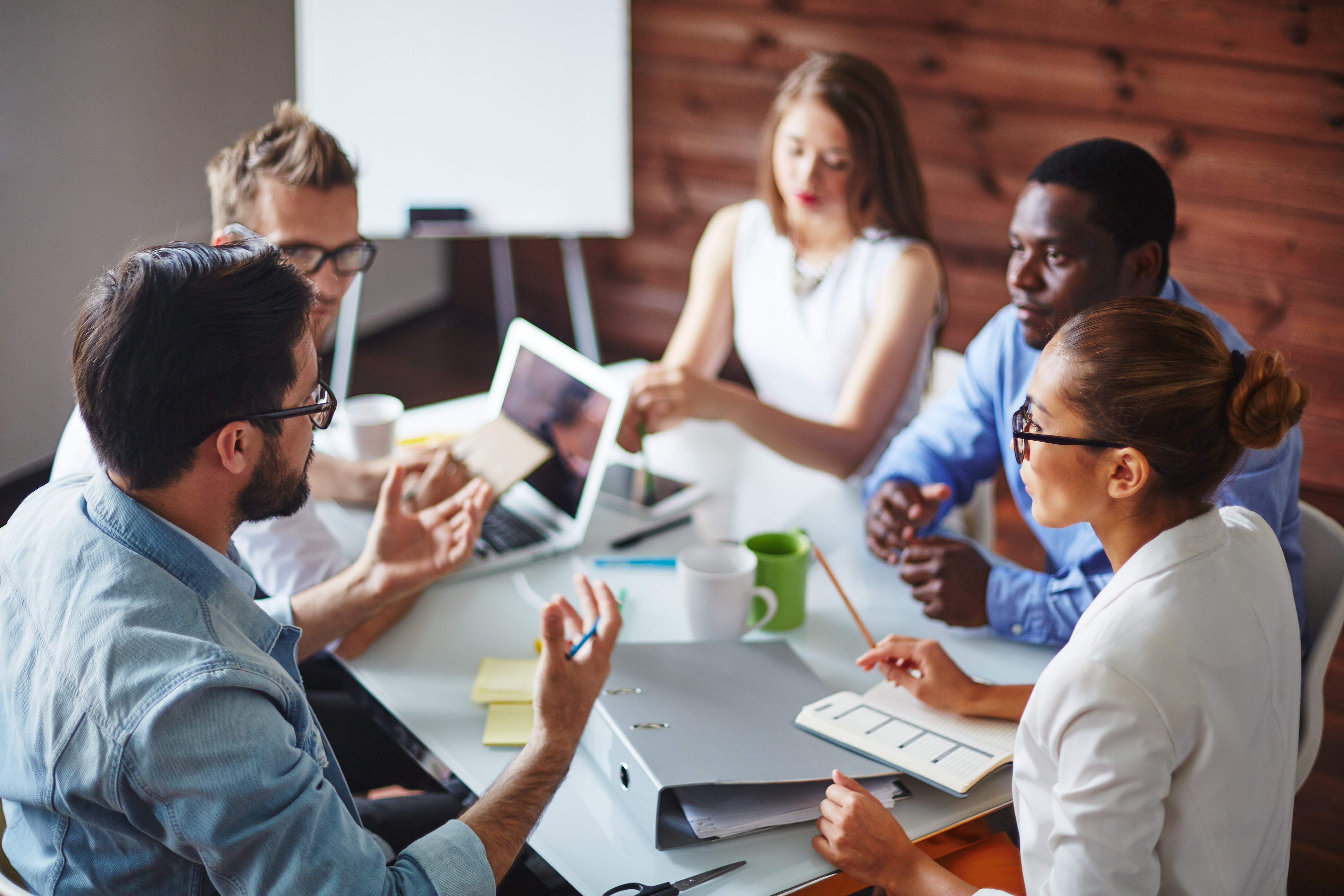 A imagem mostra um grupo de cinco pessoas em uma reunião de trabalho ao redor de uma mesa. Todos parecem estar envolvidos em uma discussão ativa, com laptops e materiais de escritório, como pastas, canecas e blocos de notas, espalhados sobre a mesa. As pessoas estão vestidas de forma casual, com roupas de cores neutras. Uma mulher à direita, vestindo um blazer branco, faz anotações enquanto escuta atentamente. Um homem de óculos, com uma camisa jeans, gesticula enquanto fala com o grupo. A atmosfera parece ser colaborativa e focada, sugerindo uma discussão sobre um projeto ou planejamento em equipe.