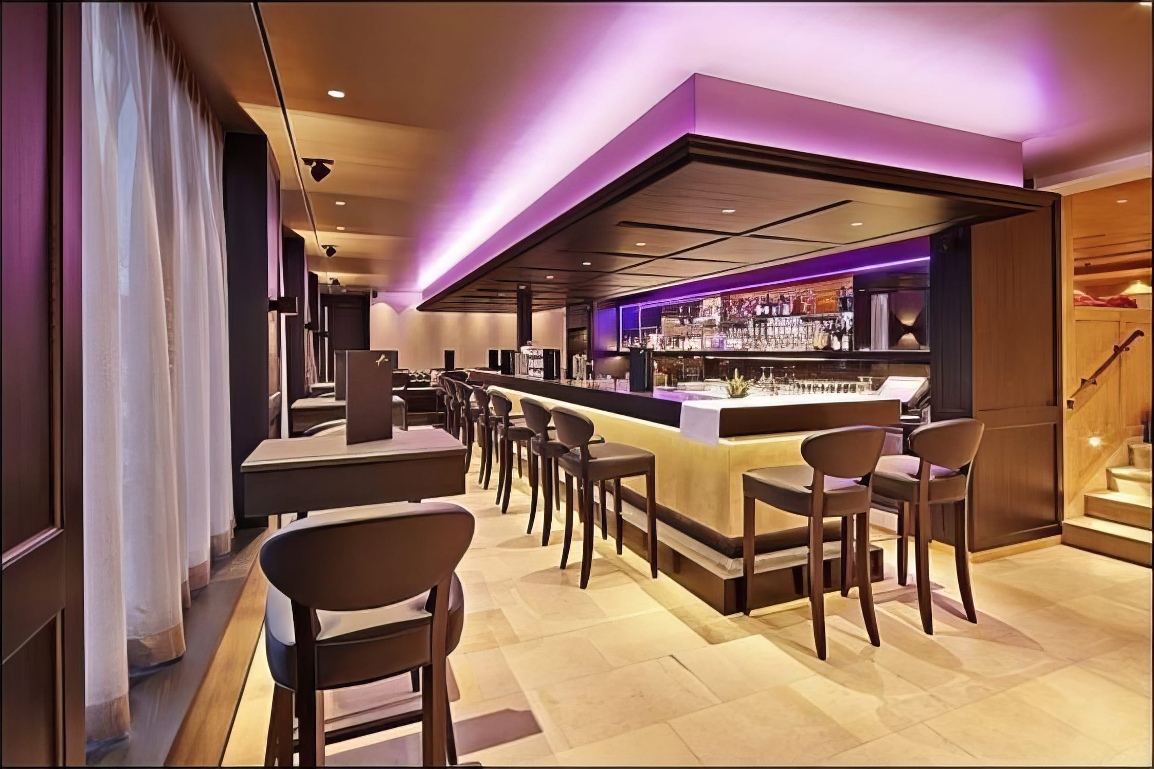 MW Interior  - Elegante Hotelbar mit indirekter violetter LED-Beleuchtung, beleuchteter Spirituosenrückwand, dunklen Holzbarhockern und cremefarbenem Natursteinboden" Beschreibung: Stimmungsvolle Hotelbar mit violettem Lichtkonzept, hinterleuchteter Spirituosenrückwand und dunklem Holzmobiliar – eine Atmosphäre, die nach dem Skitag zum Verweilen einlädt und den Abend eröffnet.