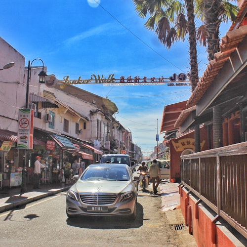 A bustling street with shops on both sides, a car driving under an archway, and a few people walking. Palm trees are visible in the background.