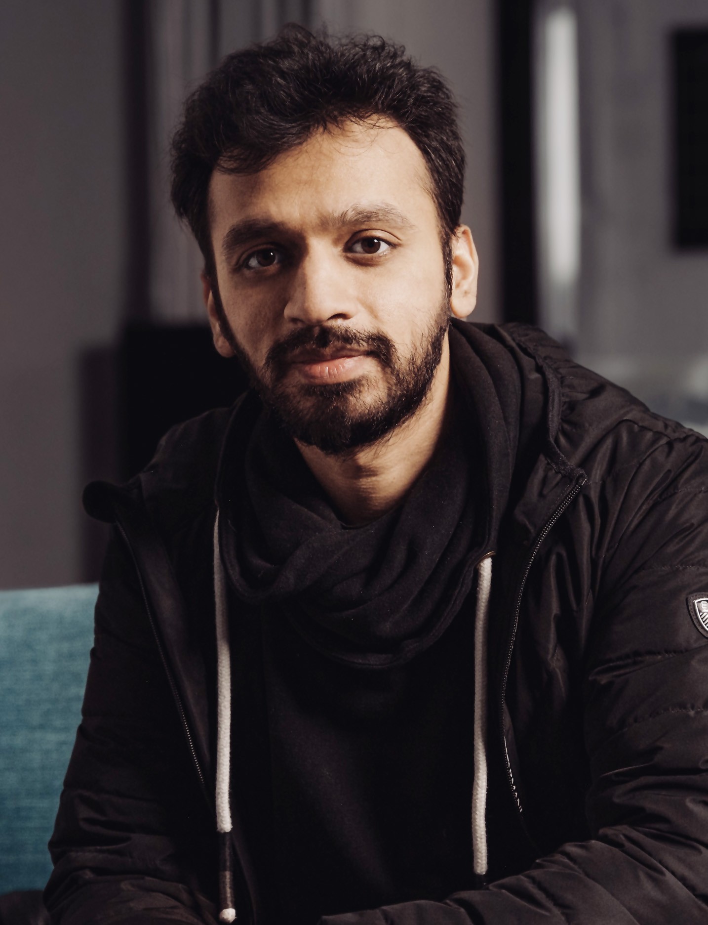 A smiling man in wearing a hoodie and black jacket sitting in an office environment.