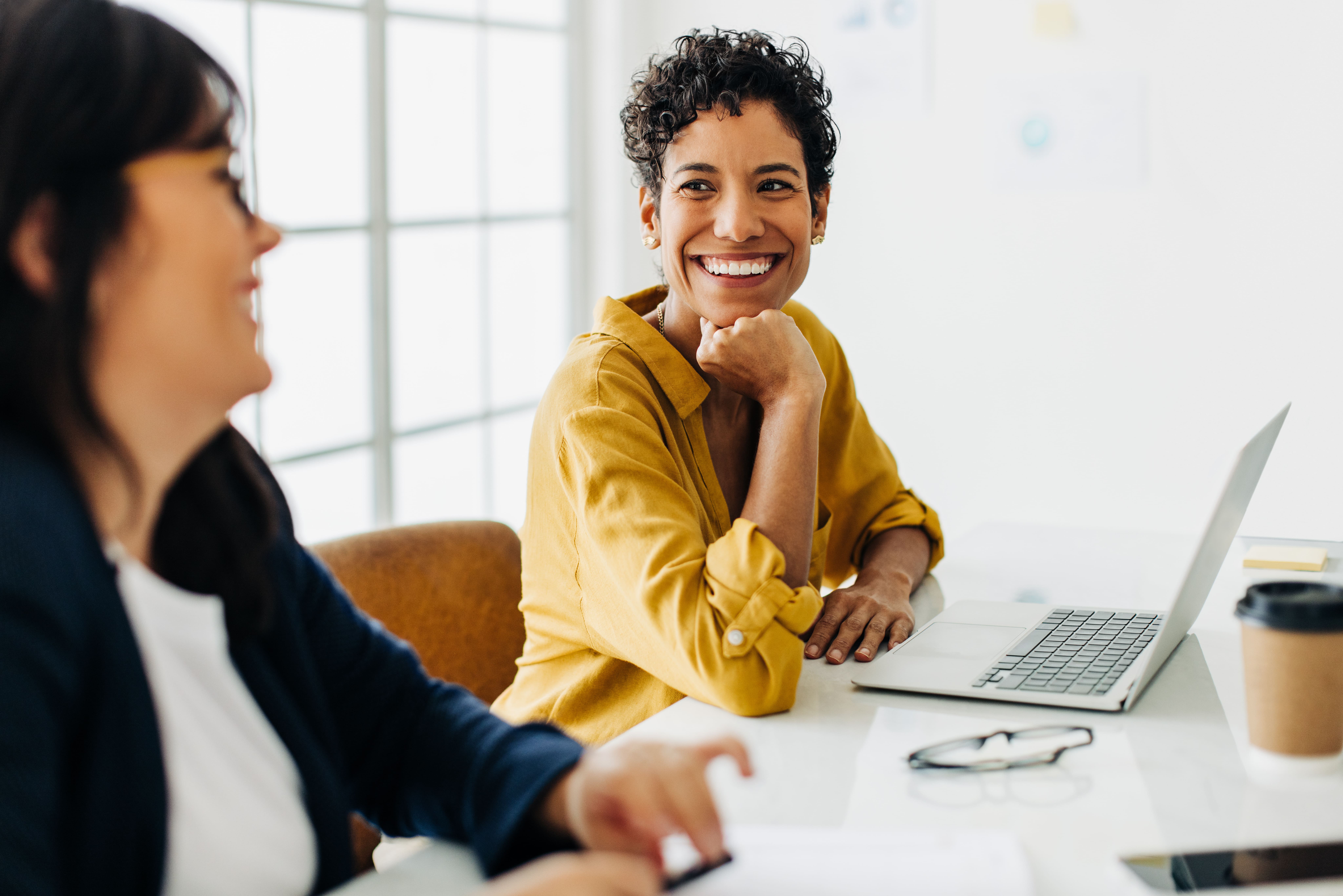 A imagem mostra duas mulheres conversando em um ambiente de trabalho, com foco em uma mulher que está sorrindo de forma calorosa enquanto olha para sua colega. Ela usa uma camisa amarela e está sentada em frente a um laptop, com uma xícara de café descartável ao lado. Seus óculos estão descansando sobre a mesa. O ambiente é bem iluminado, com luz natural entrando pelas janelas. A expressão alegre e descontraída da mulher sugere uma conversa agradável e um ambiente de trabalho positivo e colaborativo.