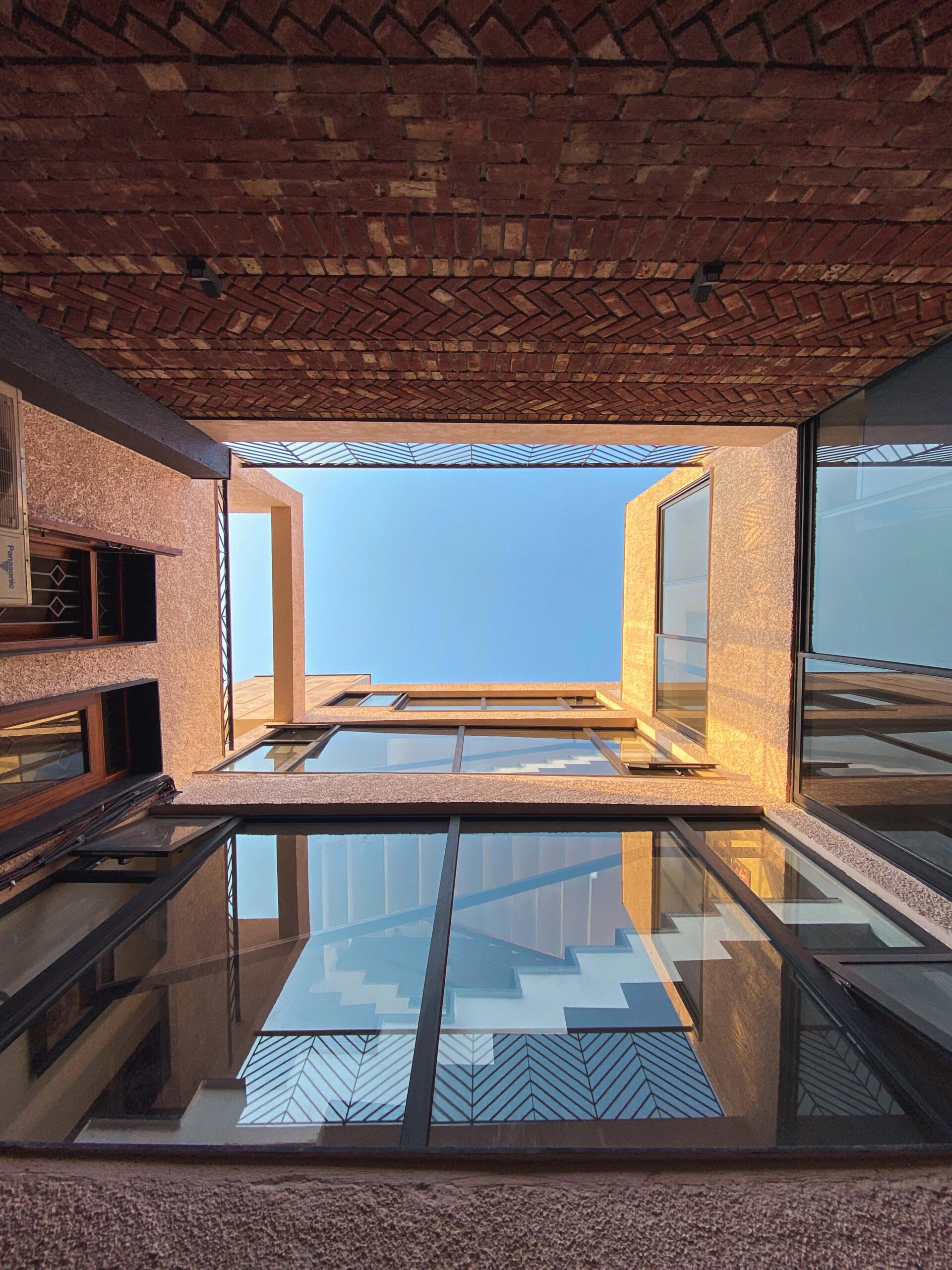 An upward view through a courtyard framed by textured walls, reflective glass, and a patterned brick ceiling opening to the blue sky.