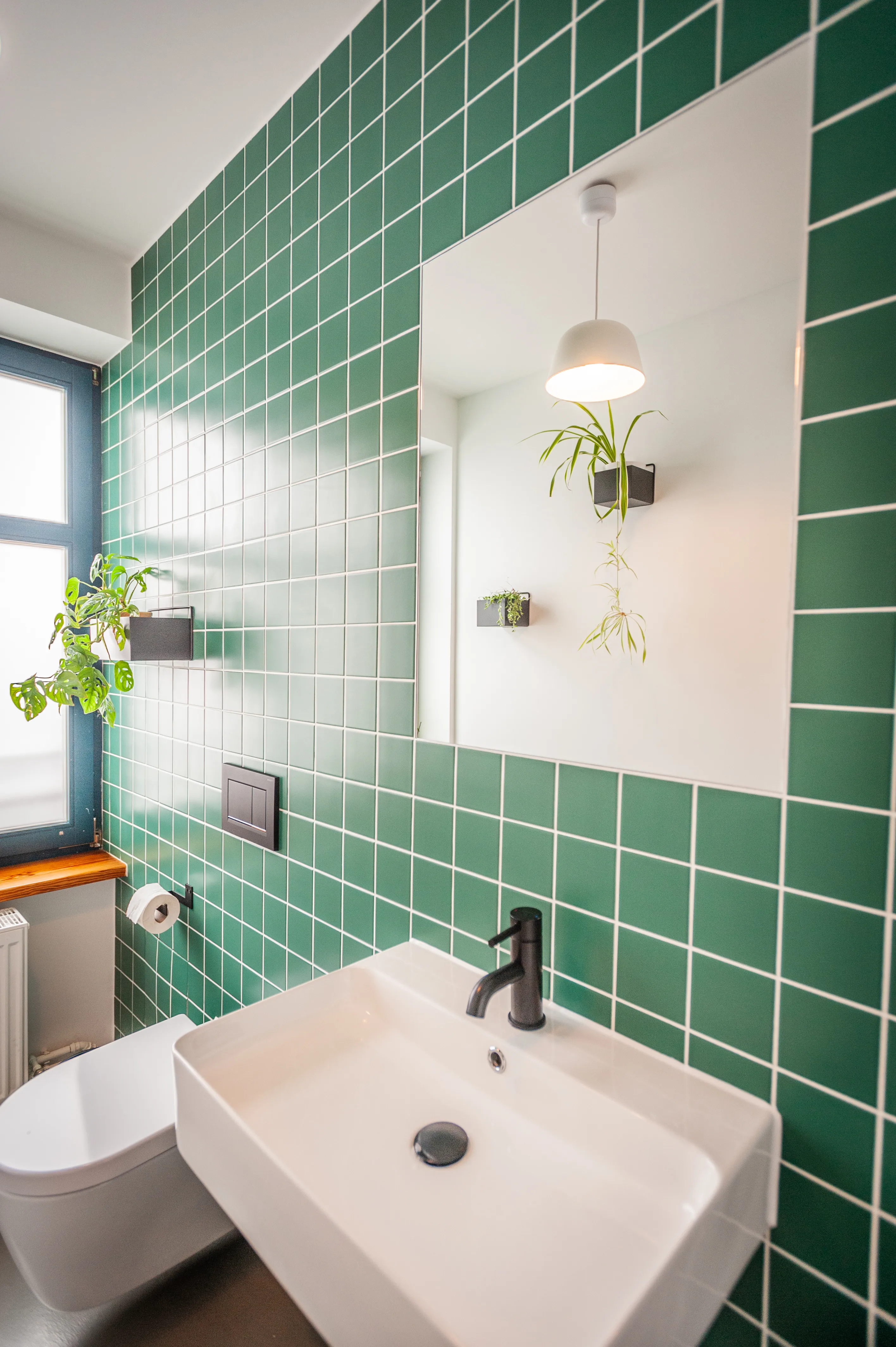 Bathroom at EHEALTH-TEC with green square tiles and white grout, a white rectangular basin with black tap, a large mirror reflecting a pendant lamp and hanging plants.