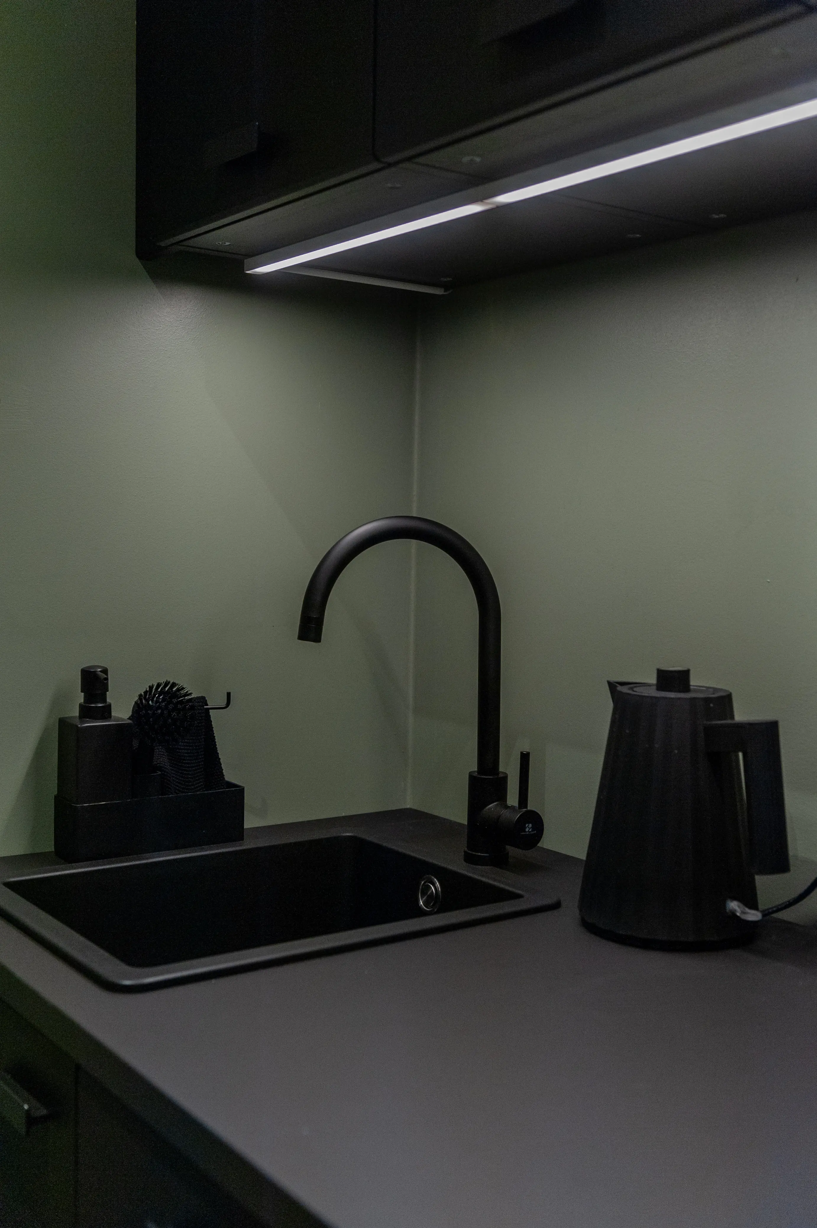 Detail of the dark kitchenette at AI START-UP BERLIN with a matte black sink and gooseneck tap, black kettle and accessories on a black counter against a sage-green wall.