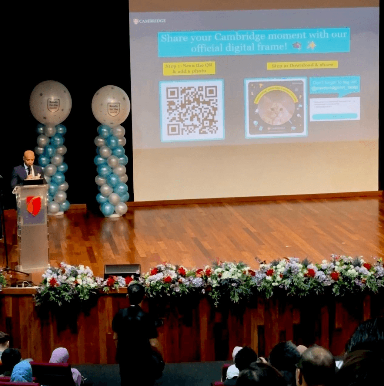 Man standing on the stage behind a podium, next to him is a screen showing a slide to promote Outstanding Cambridge Learners Awards 2026 campaign on Twibbonize.