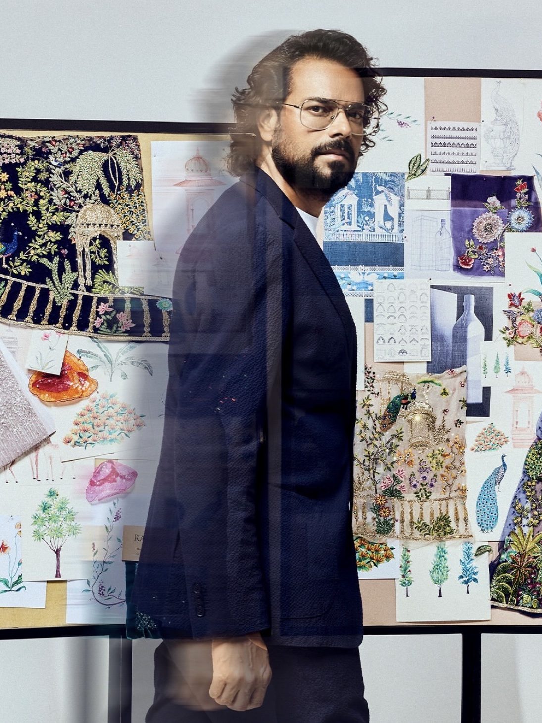 Portrait of a man standing in front of a board filled with colorful fabric swatches and sketches, showcasing intricate embroidery and design elements.