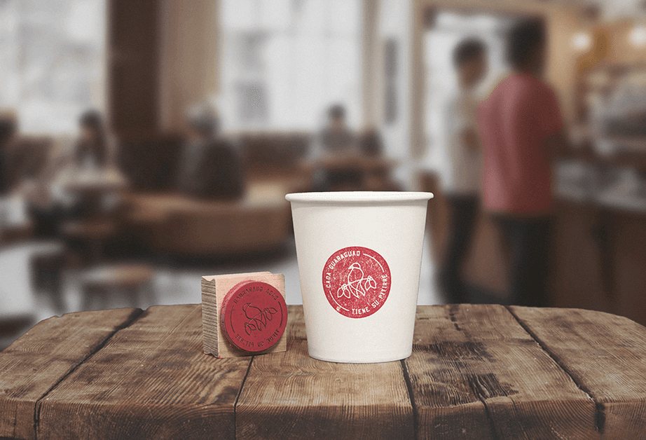 A white paper cup with a red circular logo featuring a stylized design sits on a wooden table. Beside it, a small wooden block with a red circular design is placed on the table. The background is blurred, showing a busy indoor space with people.