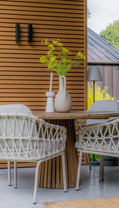 A cozy outdoor dining space with a wooden table, wrapped chairs, and a vase of yellow flowers against a modern architectural backdrop.