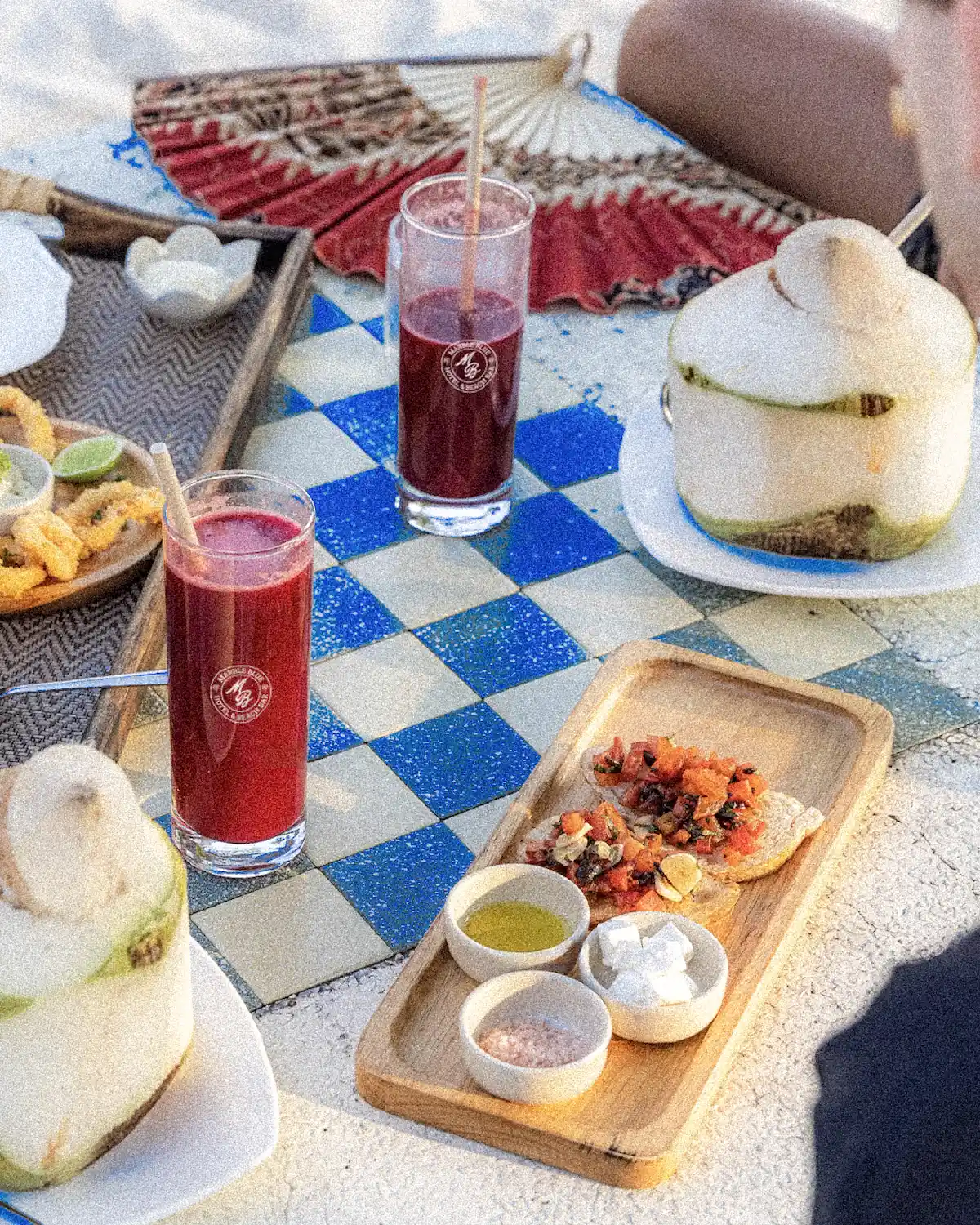 Table en carrelage damier bleu et blanc avec verres brandés Marble Blue remplis de jus rouge, noix de coco fraîche et planche bruschetta, en lumière naturelle.