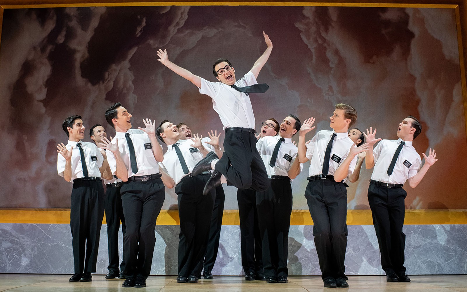 Performers in "The Book of Mormon" musical on stage, one jumping energetically.