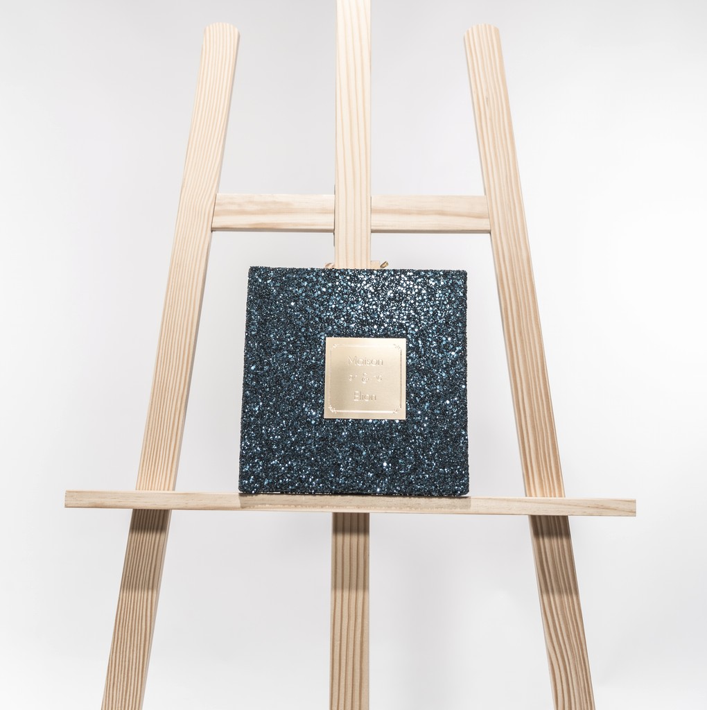 Glittering Maison Elion folio with golden plaque, poised on a minimalist wooden easel — between radiance and restraint. Folio scintillant Maison Elion orné d’une plaque dorée, posé sur un chevalet en bois minimaliste — entre éclat et retenue.