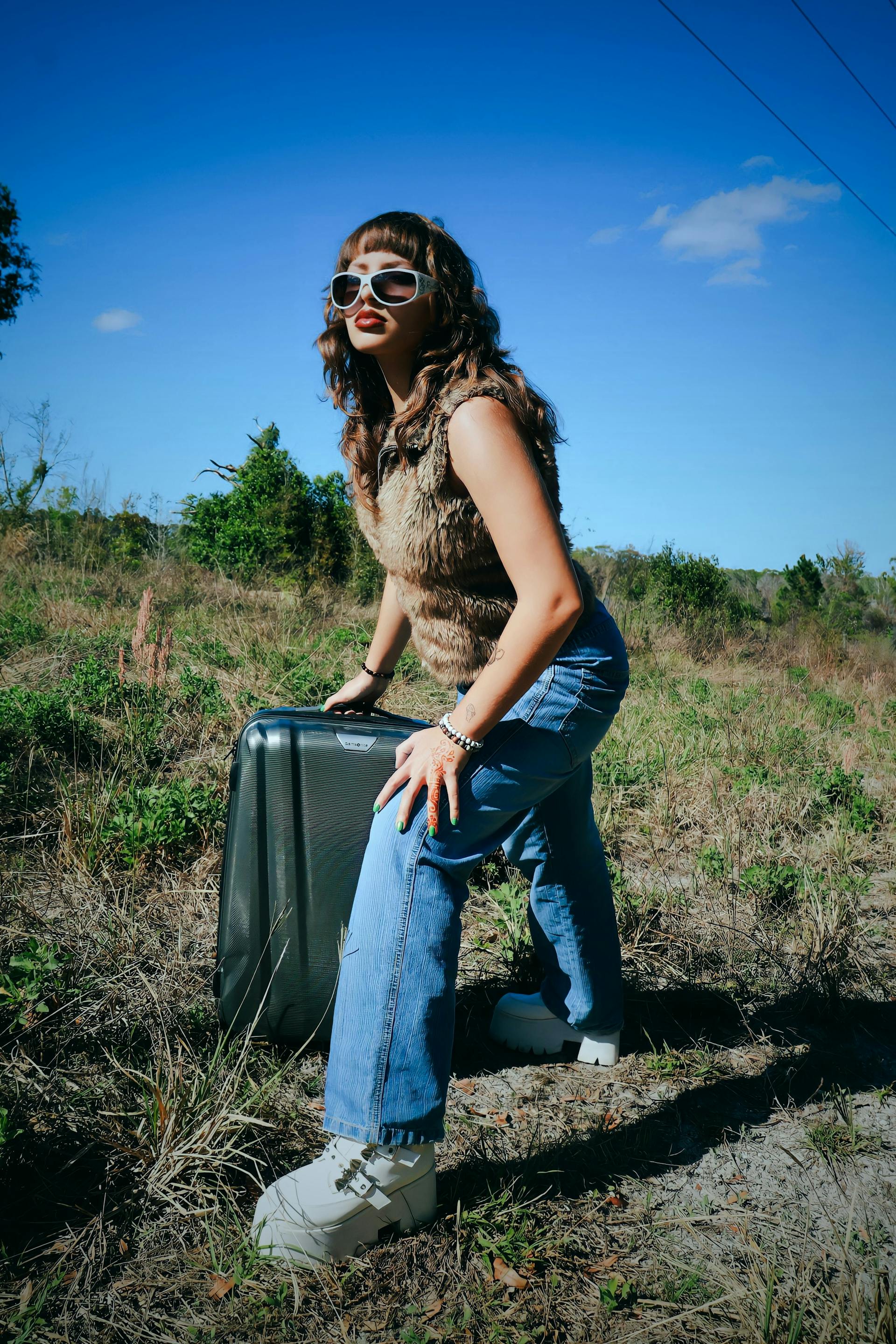 A woman in sunglasses and jeans crouches with a suitcase on a sunny, rural path