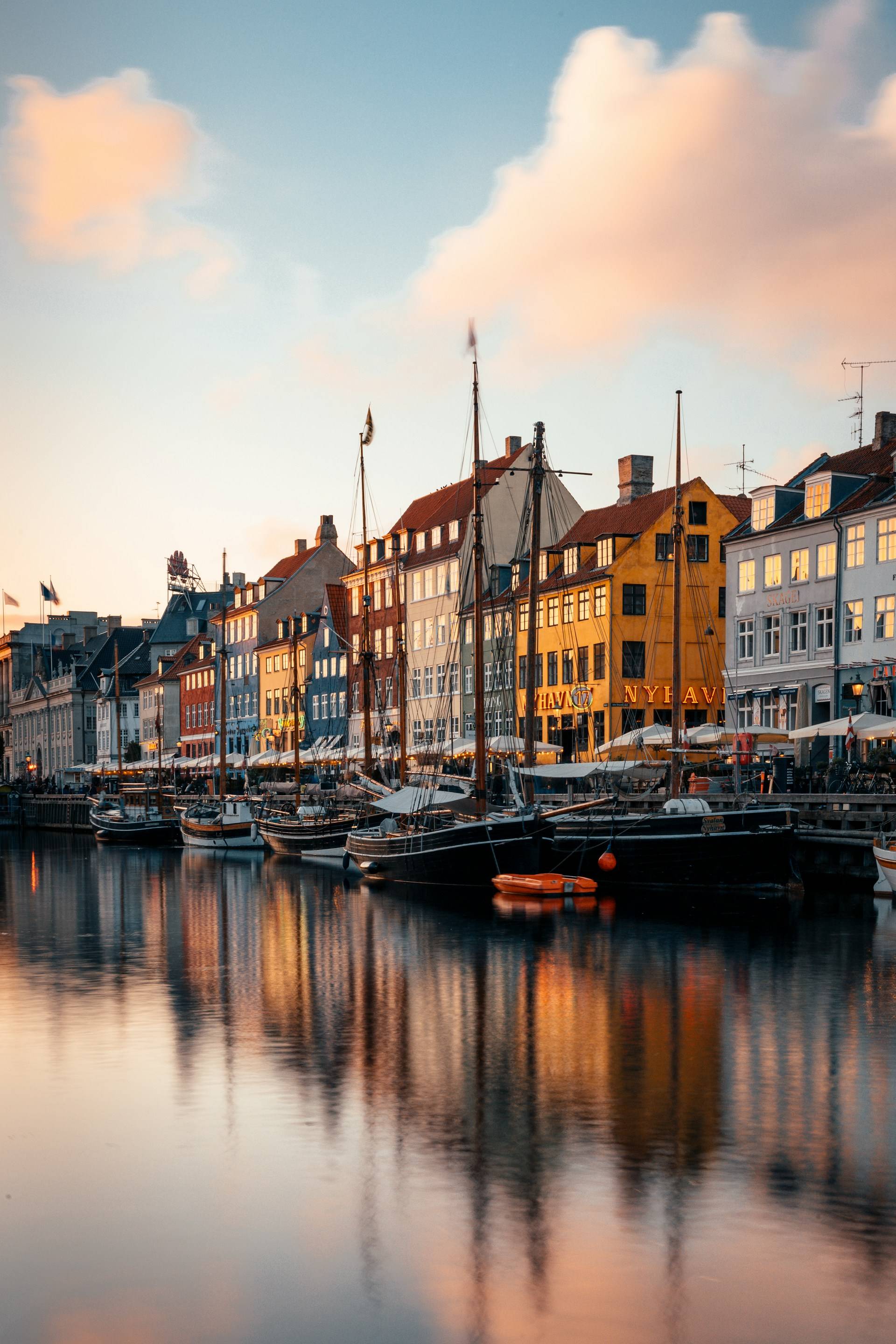 Colorful buildings and boats reflected in a calm canal at sunset, with a vibrant sky and soft clouds, creating a peaceful and picturesque atmosphere.